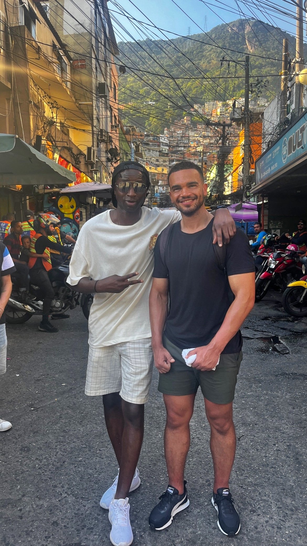 Two young men on a busy city street with tangled overhead wires, colorful buildings, and a mountain in the background. One man is wearing a white t-shirt, shorts, and sunglasses making a peace sign. The other man is wearing a black t-shirt, shorts, and sneakers, smiling with his arm around his friend's shoulder.