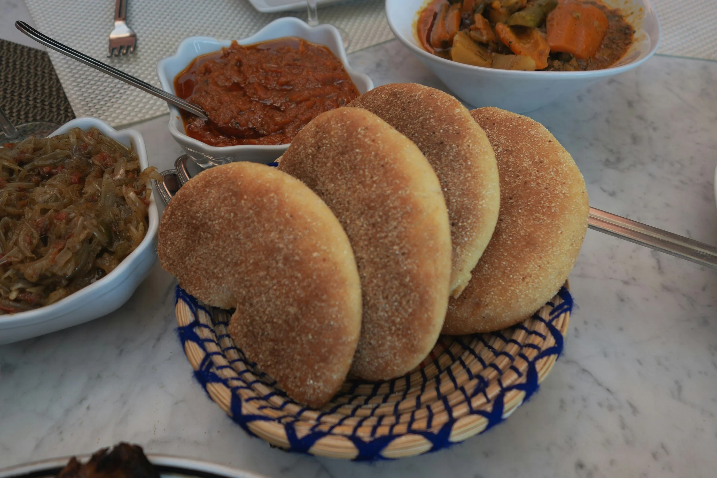 A basket of six toasted bread rolls on a marble table, with separate bowls of vegetable stew, tomato-based meat stew, and another vegetable stew in the background.