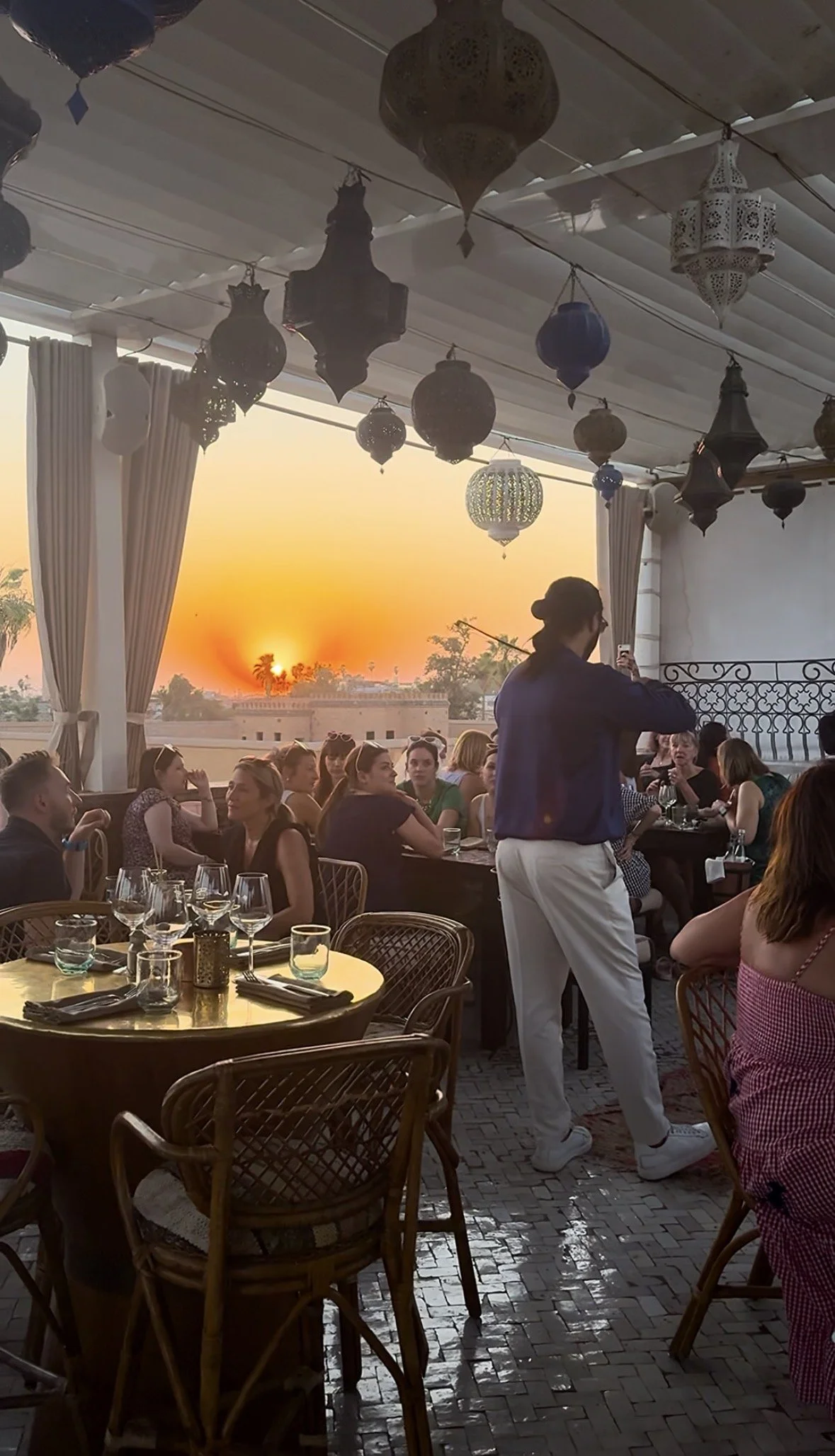 People dining at an outdoor restaurant during sunset, with hanging Moroccan-style lanterns and curtains, and a woman taking a photo or video with her phone.