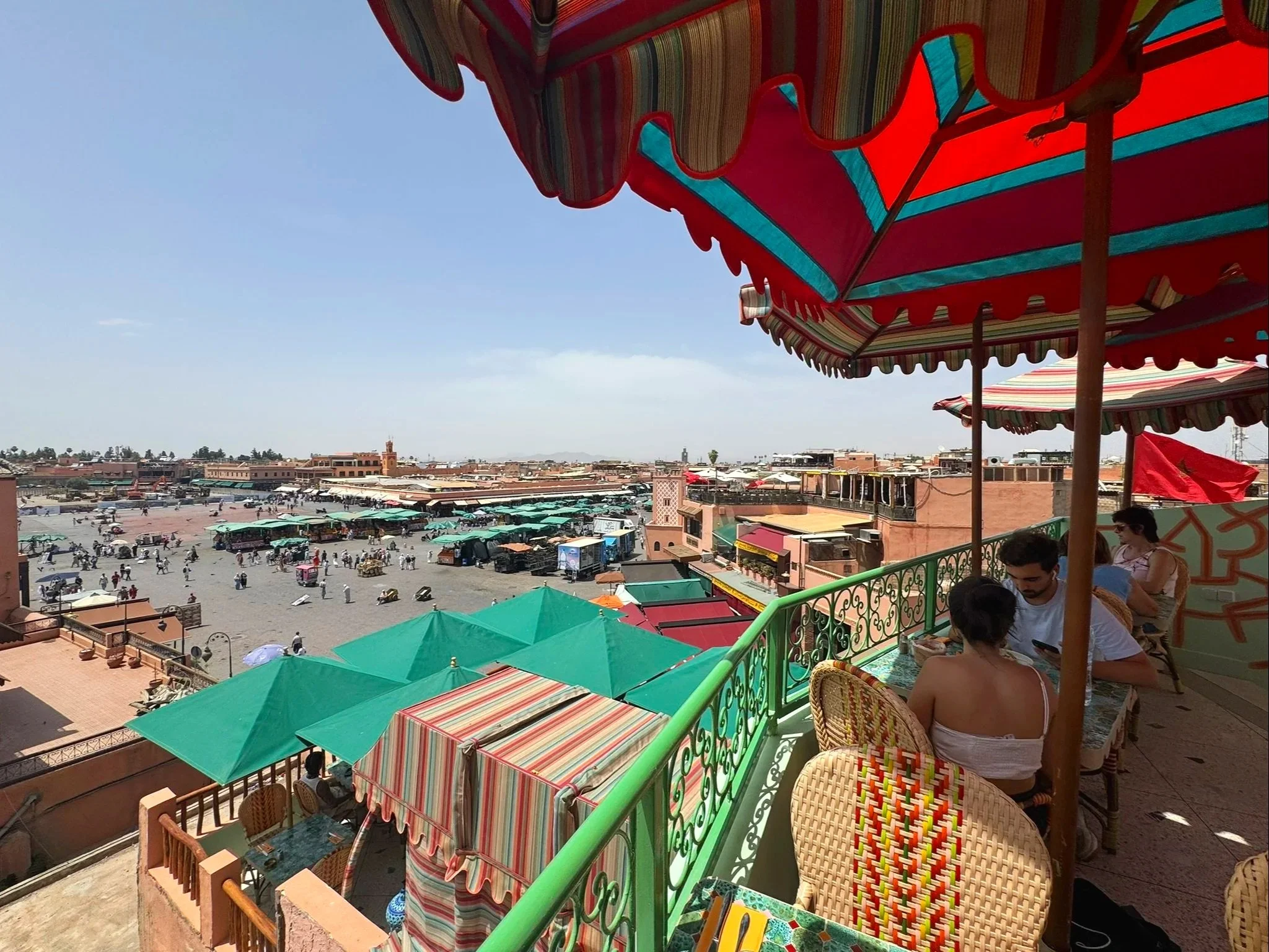 View from a balcony with colorful striped canopies overhangs, overlooking a busy marketplace or town square with many people, buildings, and green umbrellas.