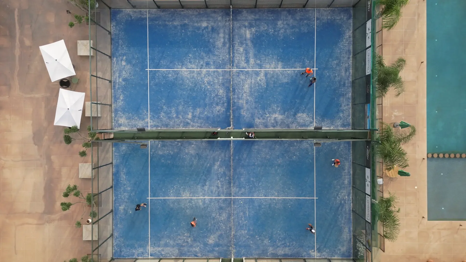 A top-down view of an outdoor tennis court with four players on the court, two on each side, and three people outside the court on the left side. The court has a blue surface with white lines, and is surrounded by a black fence, with umbrellas and potted plants along the edges.