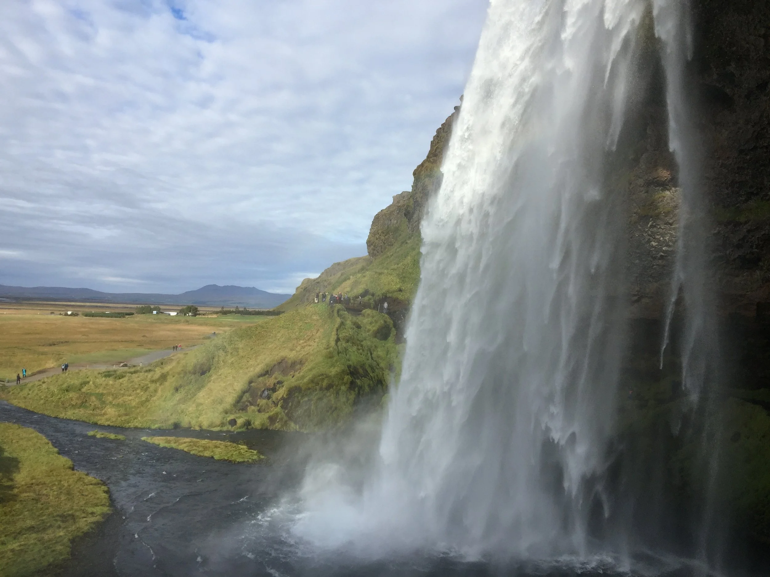 A waterfall in Iceland cascading down a cliffside with a wide river flowing at its base and a cloudy sky above, and people walking along a trail nearby.