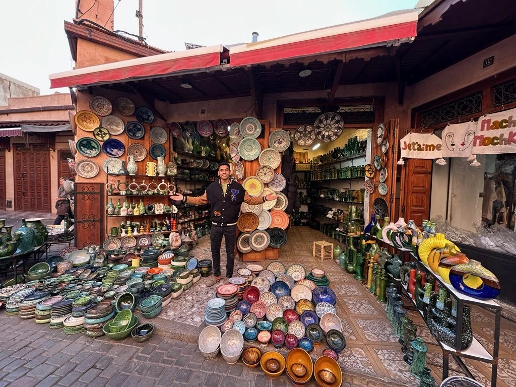 A man in Morocco stands outside his pottery shop filled with colorful handcrafted ceramic plates, bowls, and vases displayed on shelves, tables, and the ground.