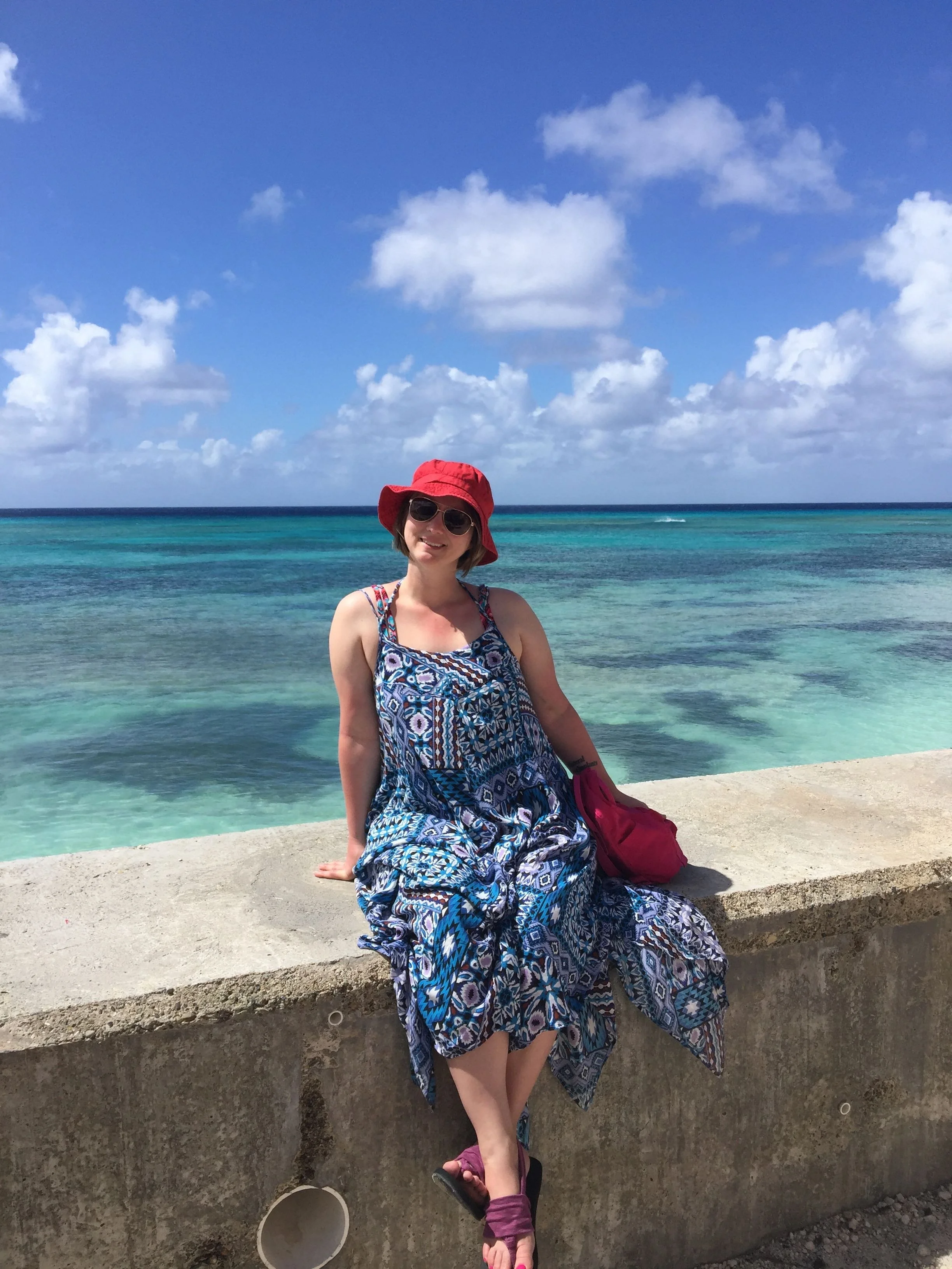 A woman sitting on a concrete ledge at the beach, smiling, wearing a red sun hat, sunglasses, a blue patterned dress, and purple sandals, with the ocean and sky with clouds in the background.