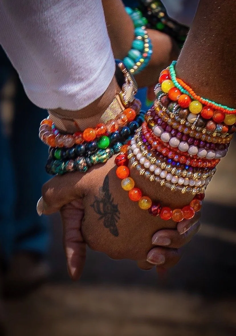Two people holding hands, adorned with colorful beaded bracelets and rings, with a tattoo of a flower on one person's hand.