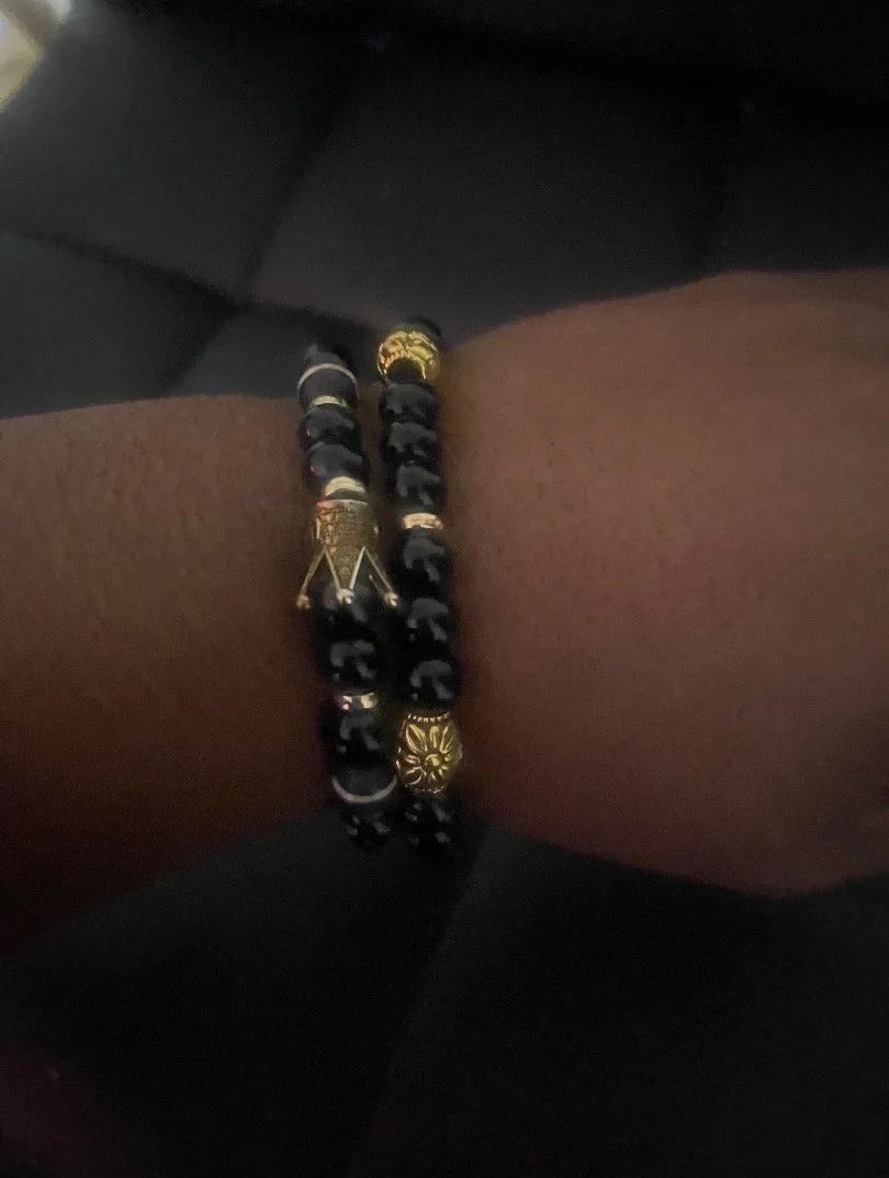 Close-up of a person's wrist wearing two black and gold beaded bracelets with decorative gold charms, against a dark background.