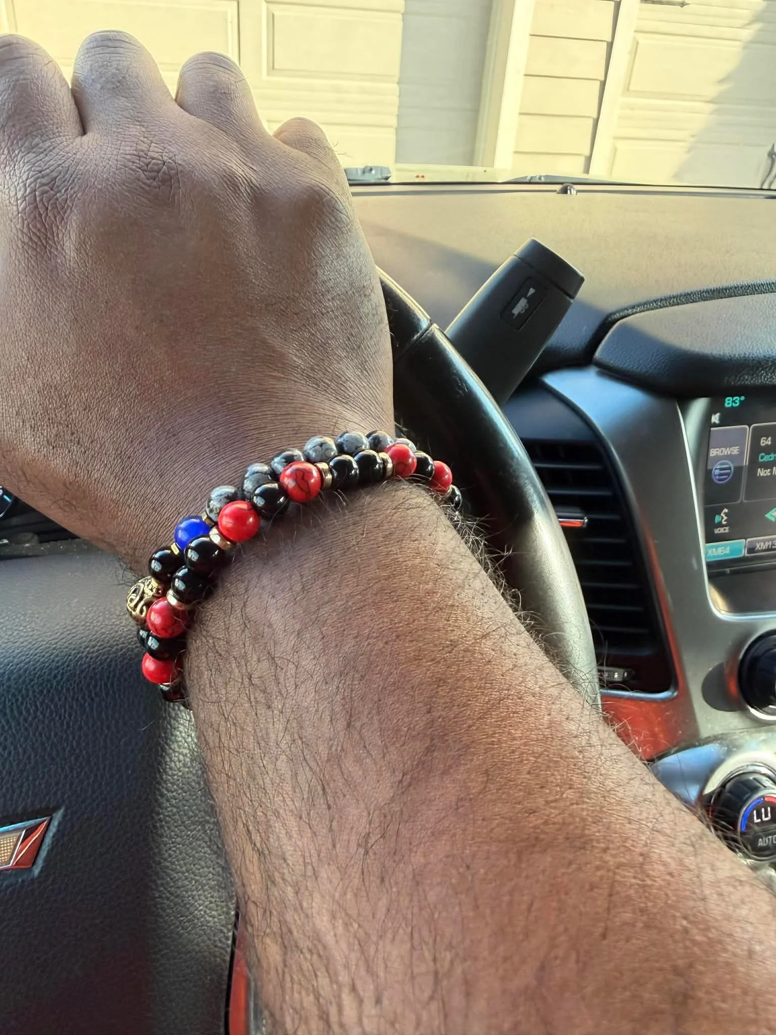 A person's arm wearing a beaded bracelet resting on the steering wheel of a vehicle. The interior of the car, dashboard, and part of the windshield are visible, with a view of a house outside.