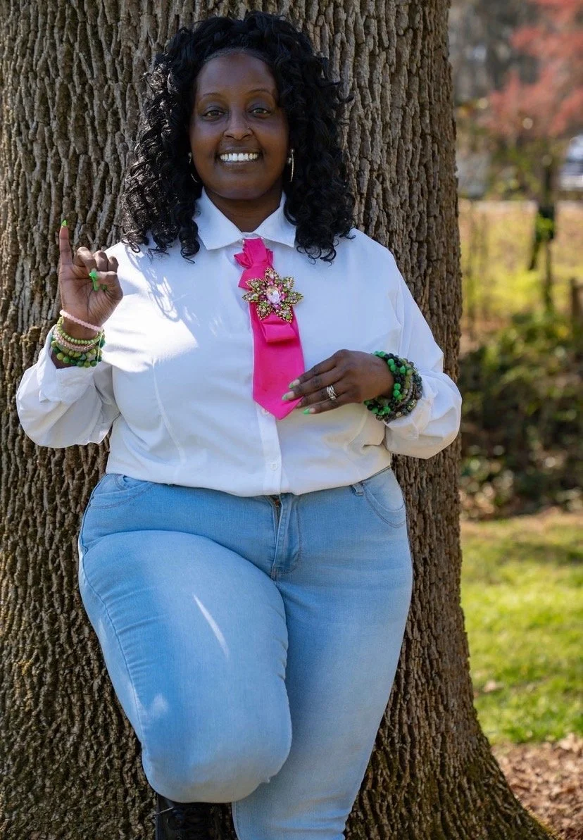 A woman standing outdoors leaning against a tree, smiling, wearing a white shirt with a pink ribbon and a decorative pin, light blue jeans, and colorful bracelets.
