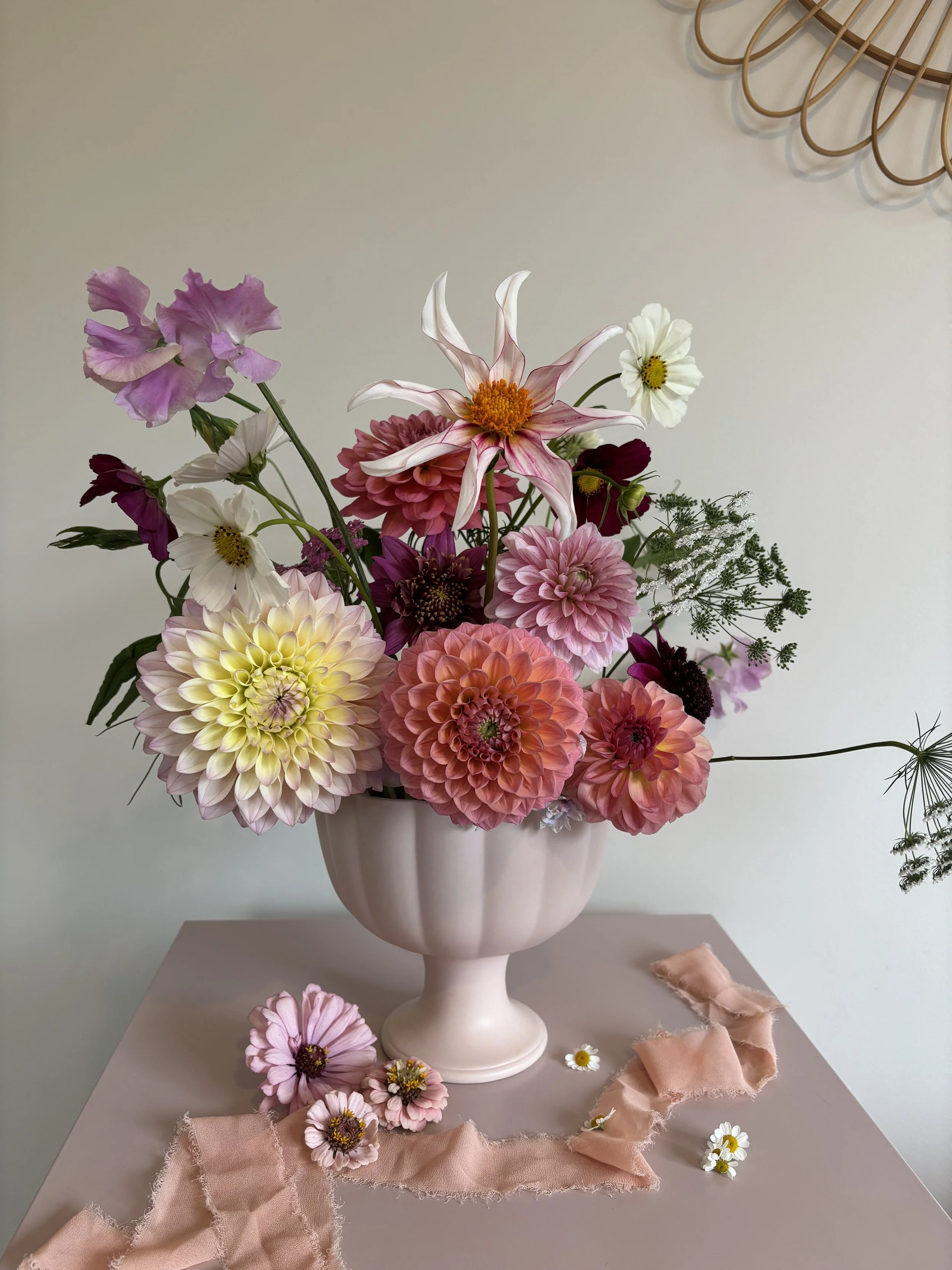 A bouquet of pink, white, and purple flowers arranged in a white vase on a table, with some flowers and pink fabric laid around the base of the vase.