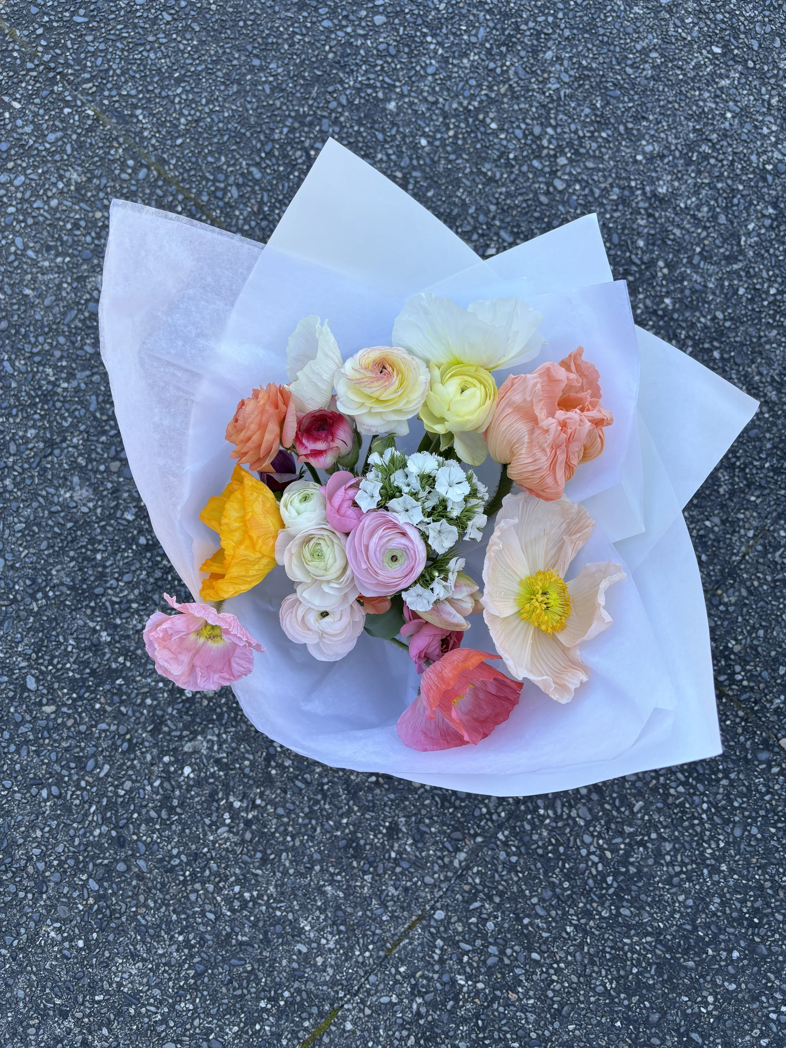 A bouquet of multicolored flowers wrapped in white tissue paper, placed on a dark, pebbled asphalt surface.