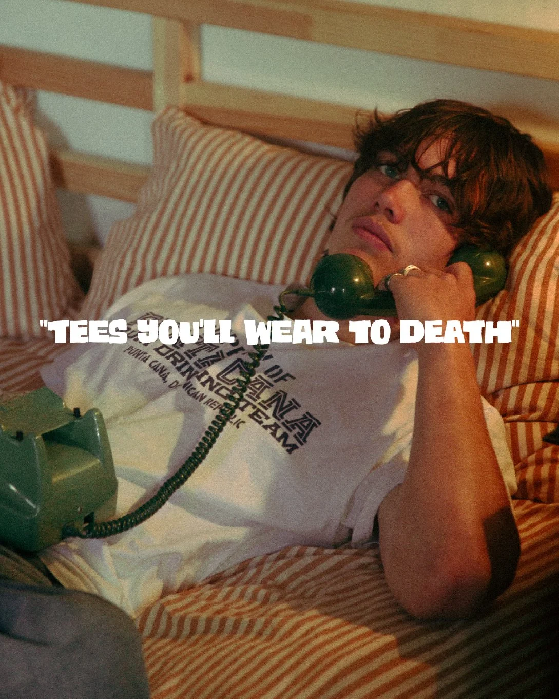 A young man with dark, messy hair lying in bed, holding a green vintage rotary phone to his ear, wearing a white T-shirt with black text, surrounded by striped pillows and sheets, with a caption that reads 'TEES YOU'LL WEAR TO DEATH'.