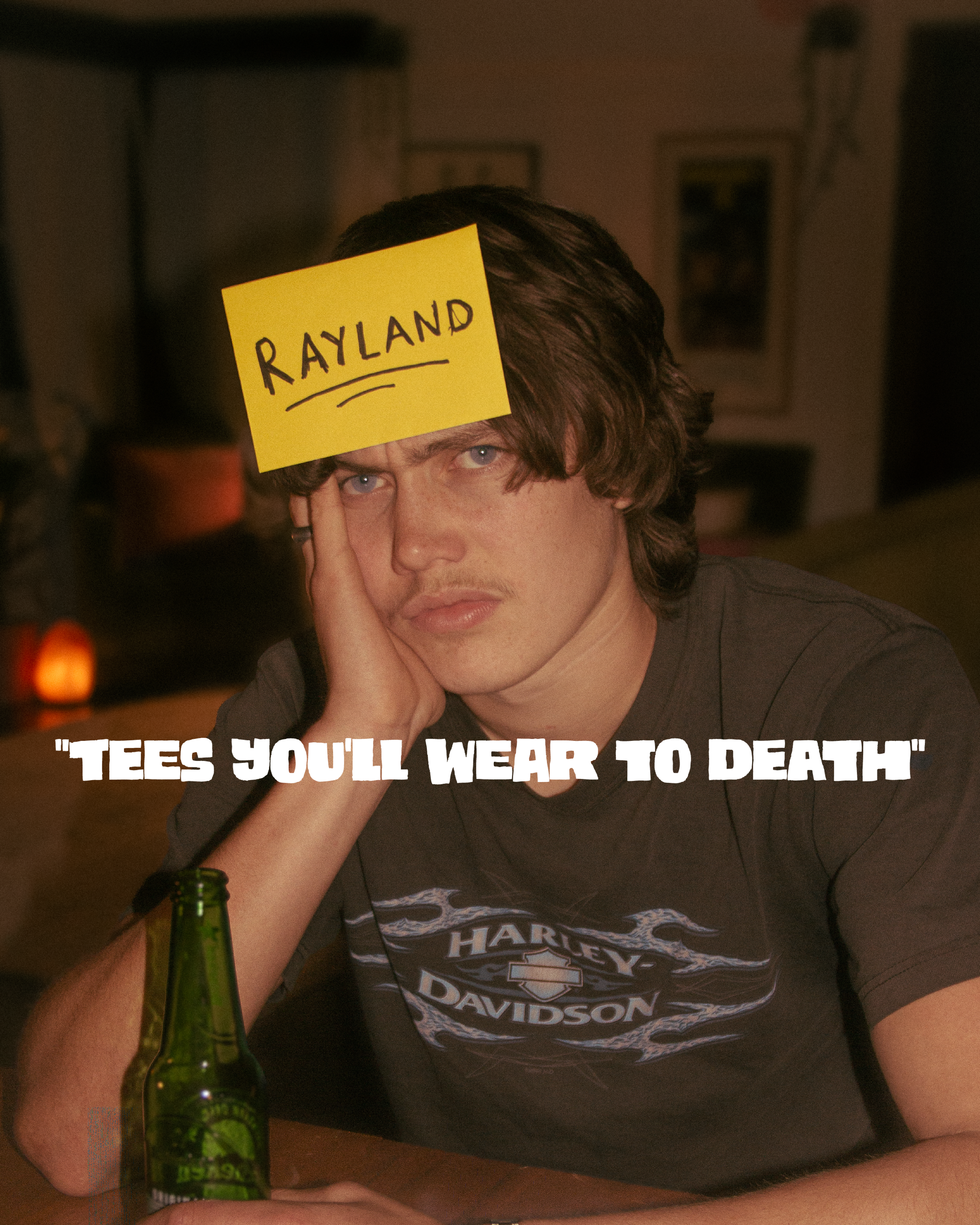 A young man with brown hair and blue eyes sitting at a table, wearing a gray Harley Davidson T-shirt, with a yellow post-it note on his forehead that reads "RAYLAND." In front of him is a green glass beer bottle. The background appears to be a dark-c