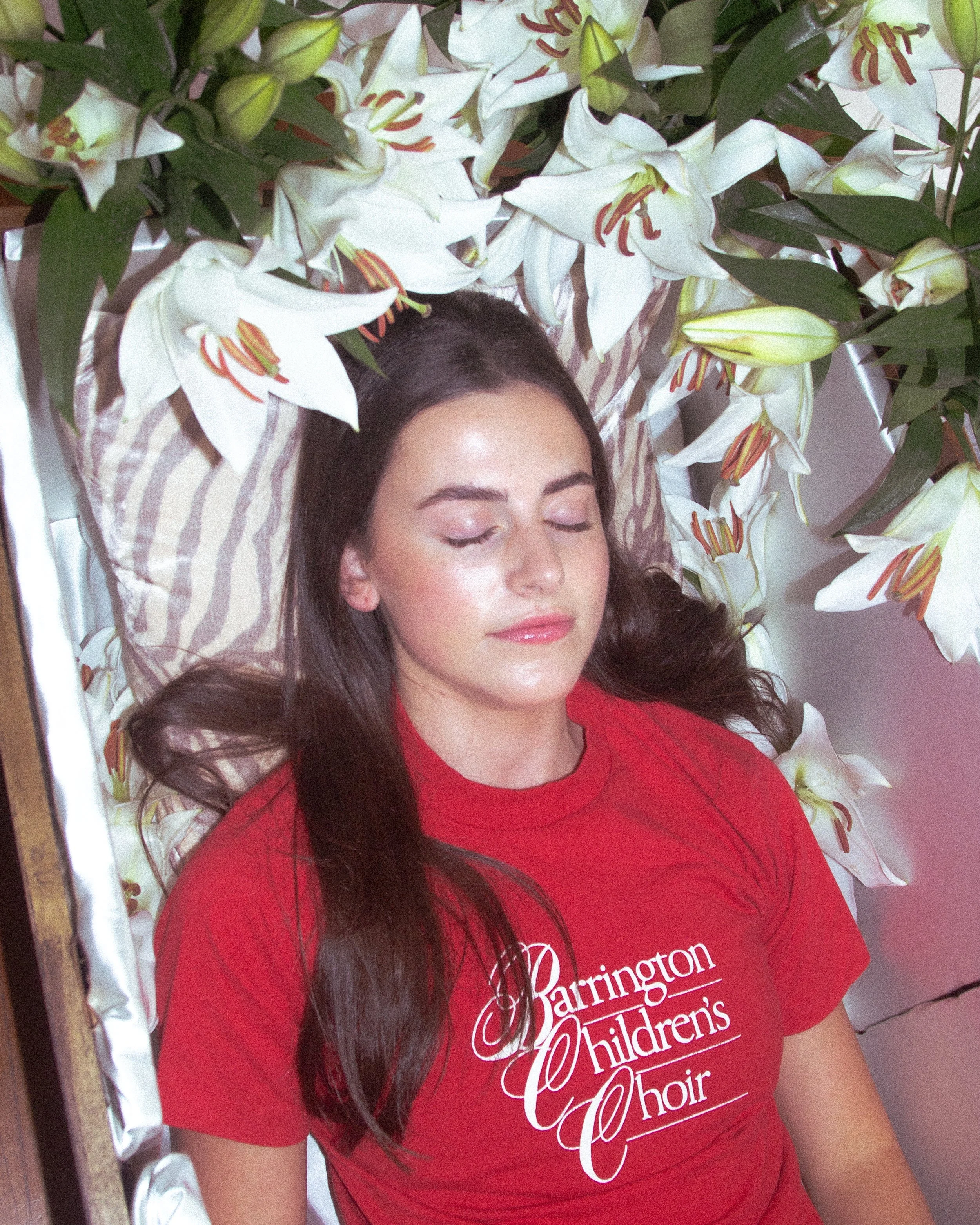 A woman with closed eyes lying on a bed surrounded by white lilies with orange and yellow stamens.