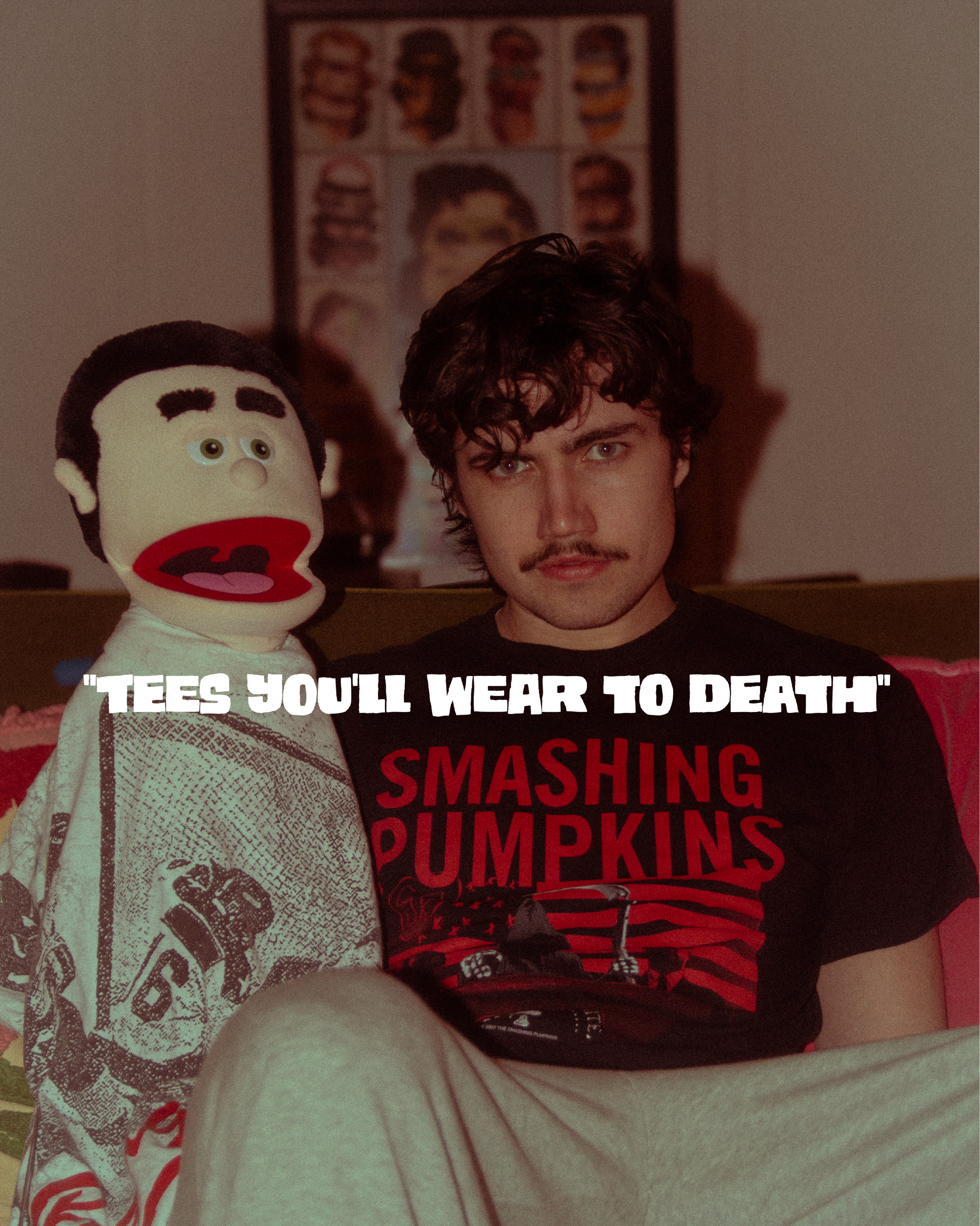 A young man with curly dark hair and a goatee sitting on a bed wearing a black and red T-shirt that has a phrase about smashing pumpkins. To his left, there is a puppet with a large head, black hair, cream-colored face, and red lips, with Chinese cha