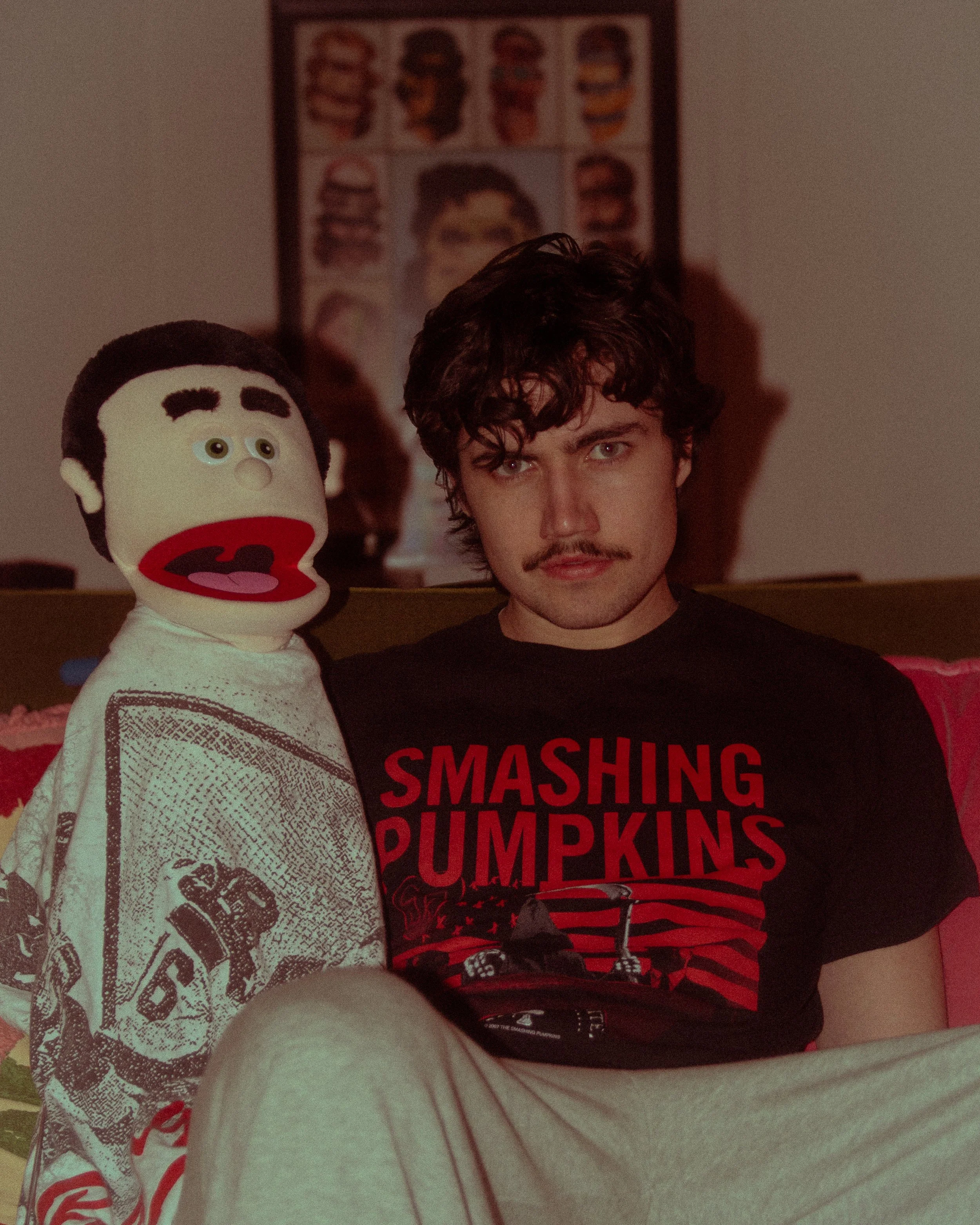 A young man with dark, curly hair and facial hair is sitting on a bed, wearing a black t-shirt with red text that reads 'SMASHING PUMPKINS'. Behind him, there is a poster featuring various styles of a person's hair, and a plush toy of a cartoon character with a white face, black hair, large brown eyes, and an open mouth with a red tongue.