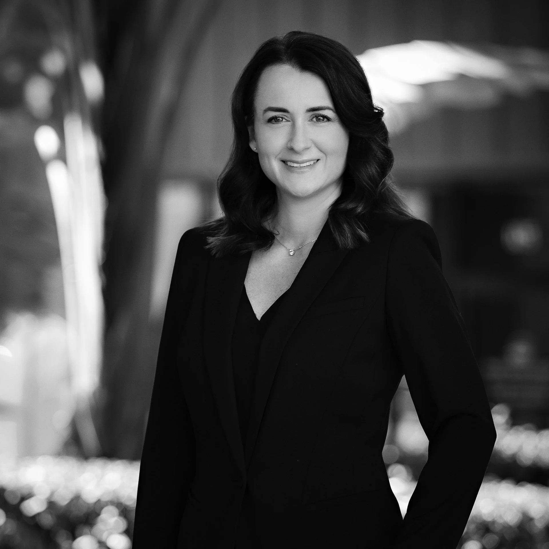 Black and white portrait of a woman with shoulder-length wavy hair, wearing a blazer, smiling, with a bokeh background.