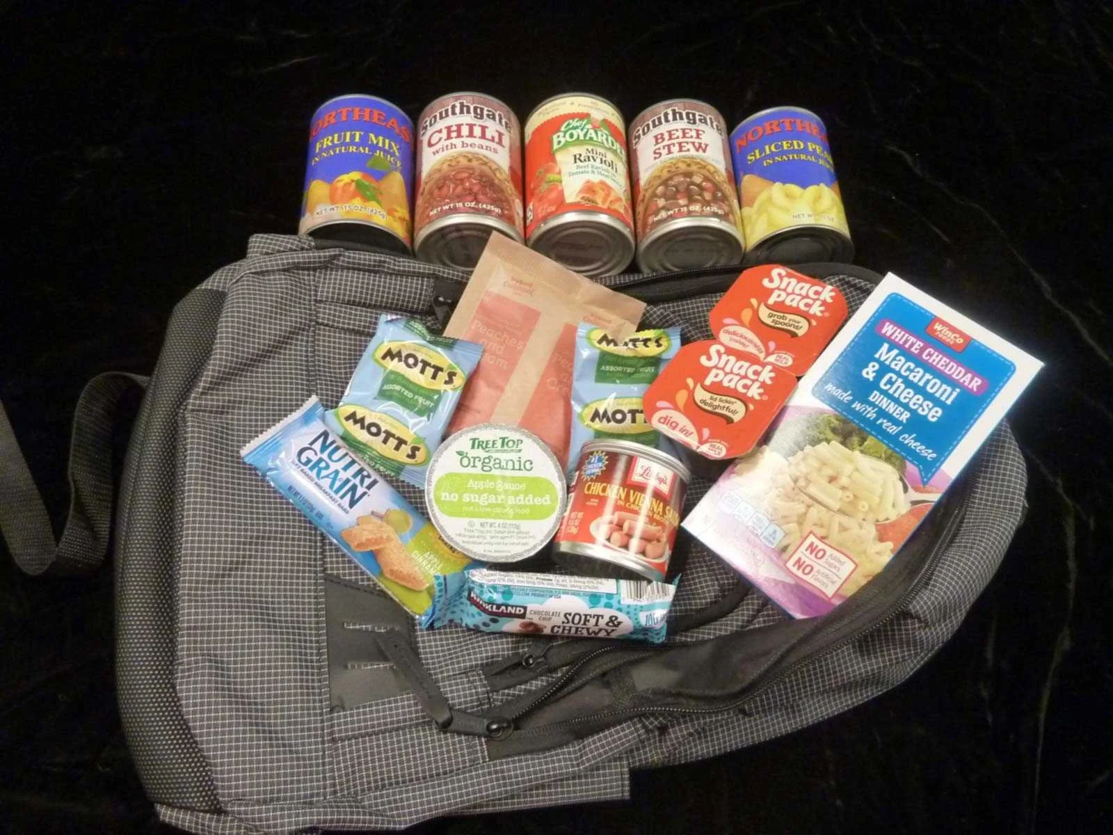 A gray backpack on a dark surface with various food items and canned goods placed on top. The food items include instant soup cans, snack packs, cheese and macaroni dinner, apple sauce, snack bars, and bagged trail mix.