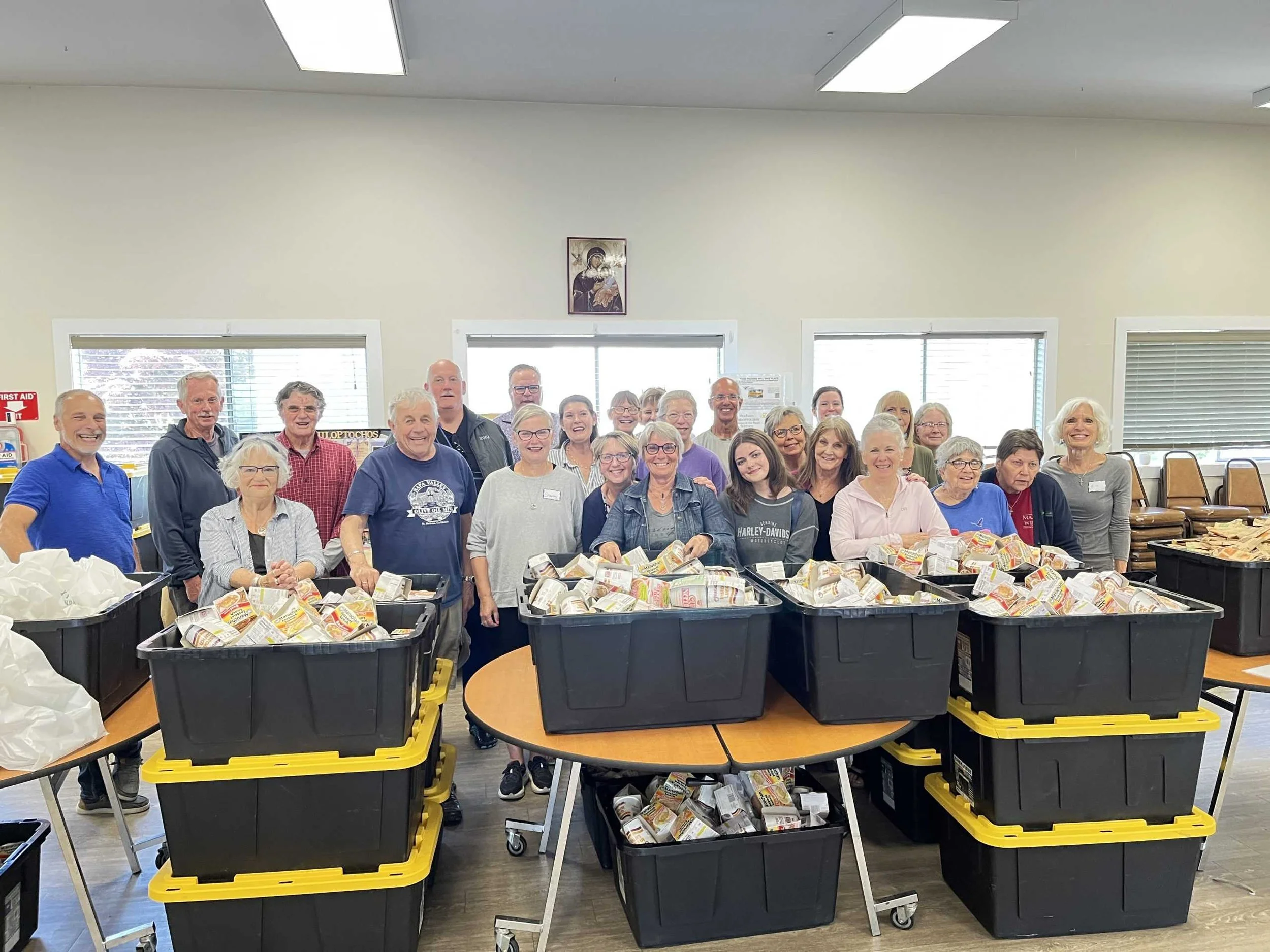Group of people standing behind tables with large black bins filled with packaged food, in a room with windows, ceiling lights, and a religious icon on the wall.