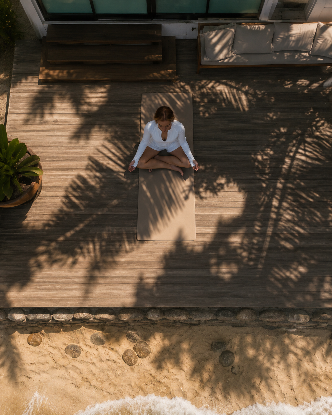 A woman practicing yoga in a seated meditation pose on a mat on a wooden deck, with shadows of palm leaves overhead and beach sand with stones and water at the bottom of the image.