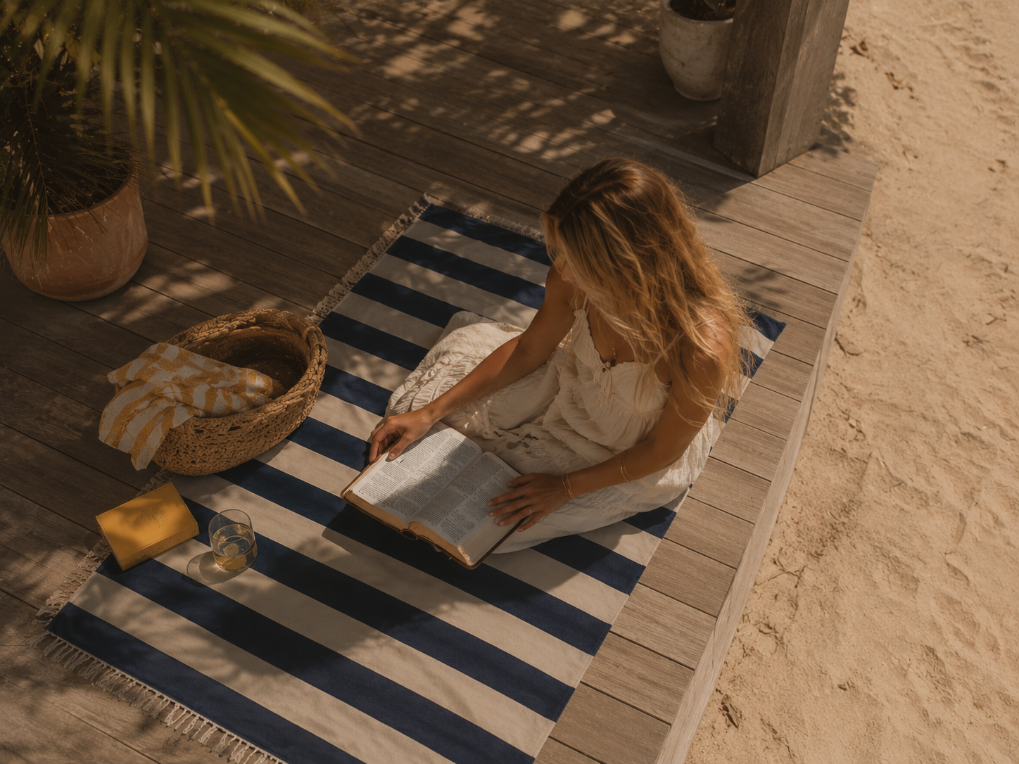 A woman with long blonde hair, wearing a white dress, sits on a blue and white striped rug on a wooden deck. She is reading a large open book, with a glass of drink, a yellow box, and a woven basket nearby. There are potted plants and sandy ground around her, with shadows from the plants and structure.