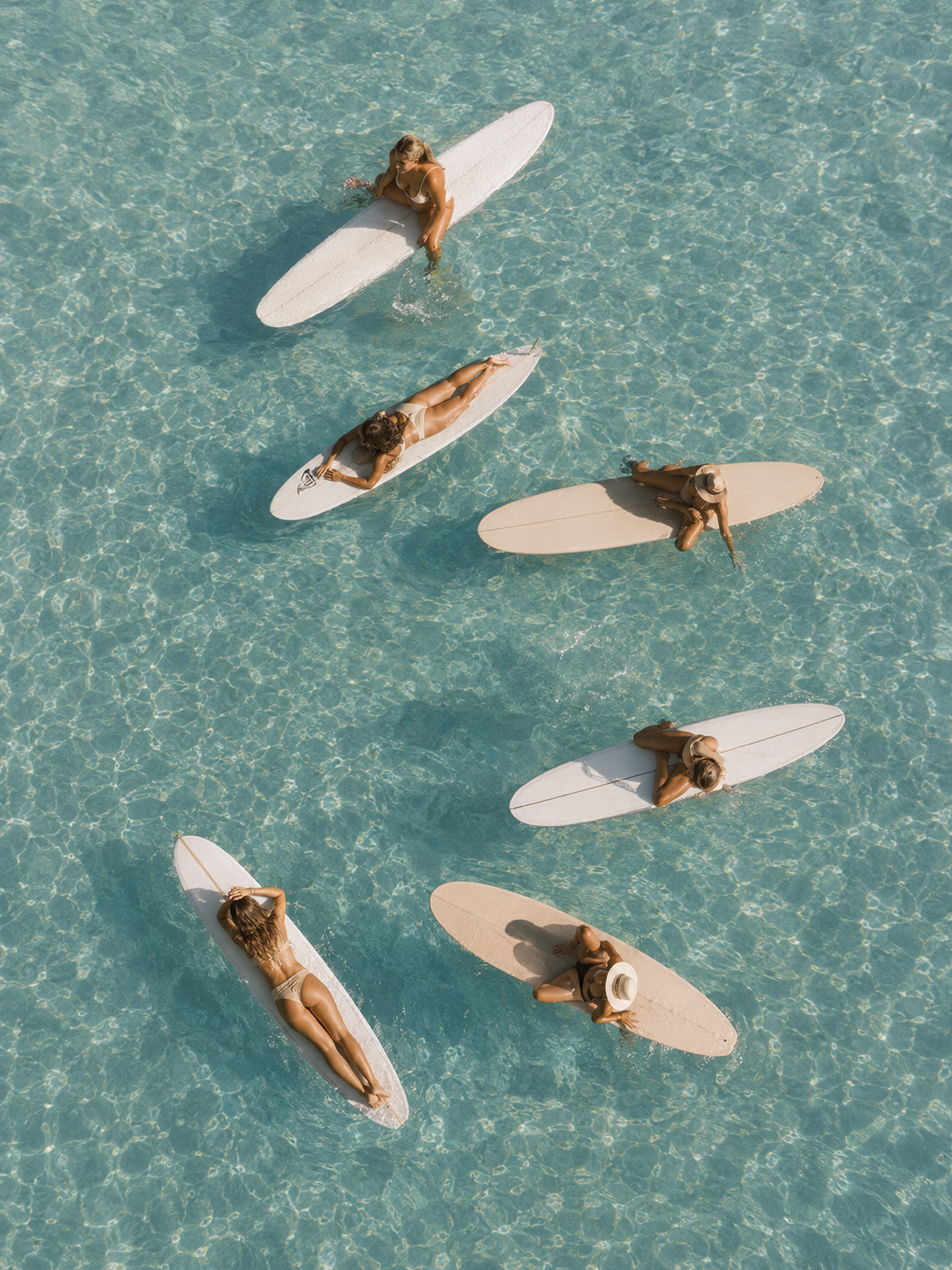 Six women in swimsuits and sun hats relaxing on paddleboards in clear turquoise water.