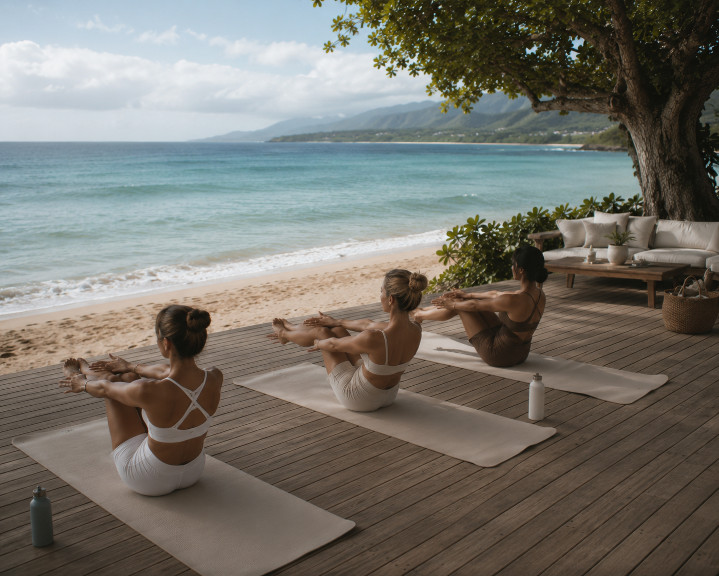 Three women practicing yoga on a wooden deck overlooking the beach and ocean. They are seated in a boat pose on yoga mats with water bottles nearby, under a tree with outdoor furniture nearby.