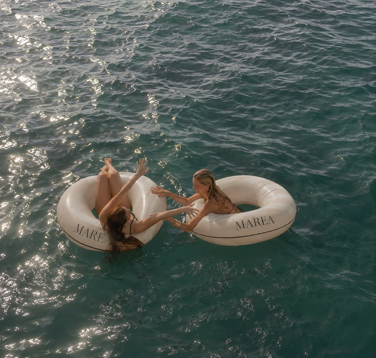 Two women in swimsuits enjoying a playful moment on life rings in the water, holding hands and smiling, with the sun reflecting on the waves.