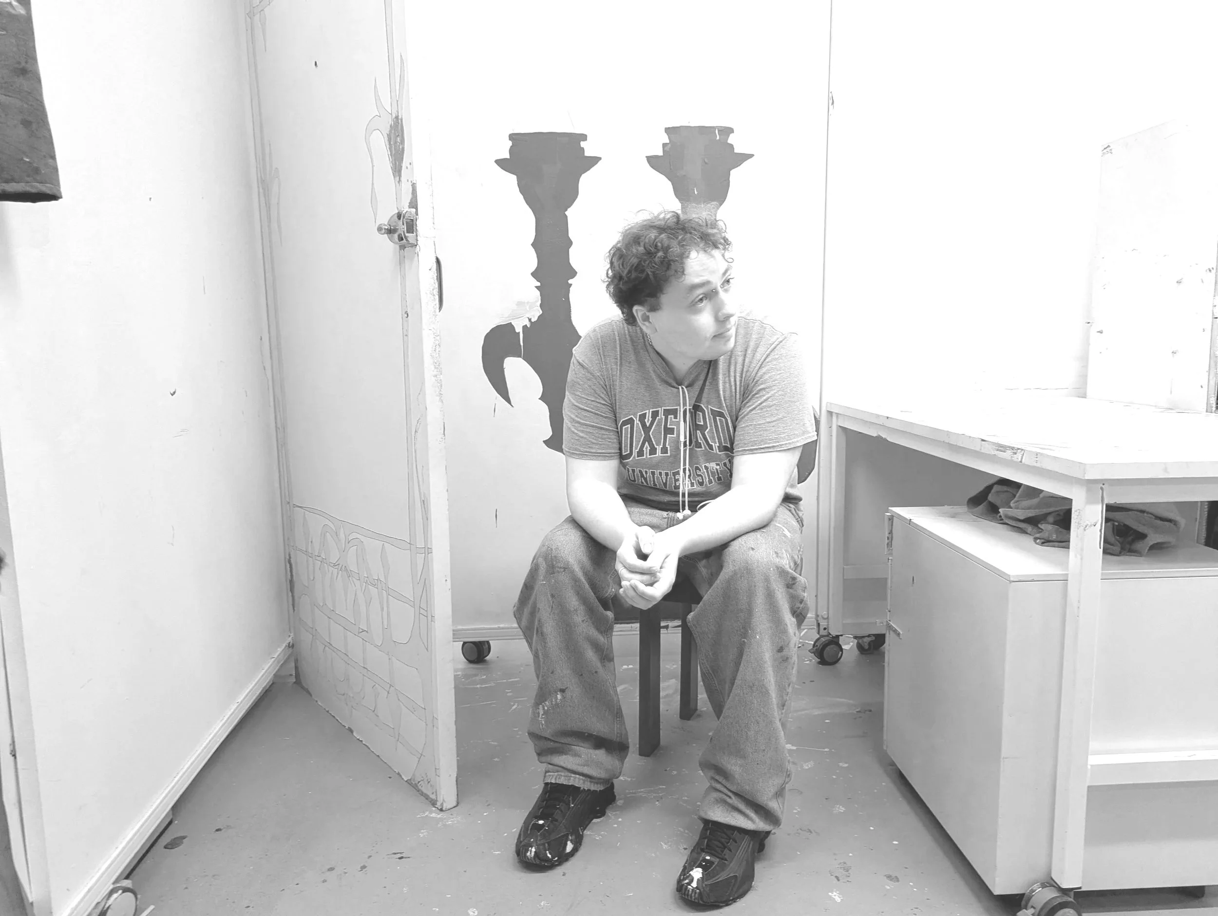 A young man with curly hair, wearing a gray Oxford University T-shirt, sitting on a stool in an art studio. The studio has white walls, one decorated with black silhouette cutouts of candlesticks. There is a ladder and a wooden table with art supplies and clothes on it.