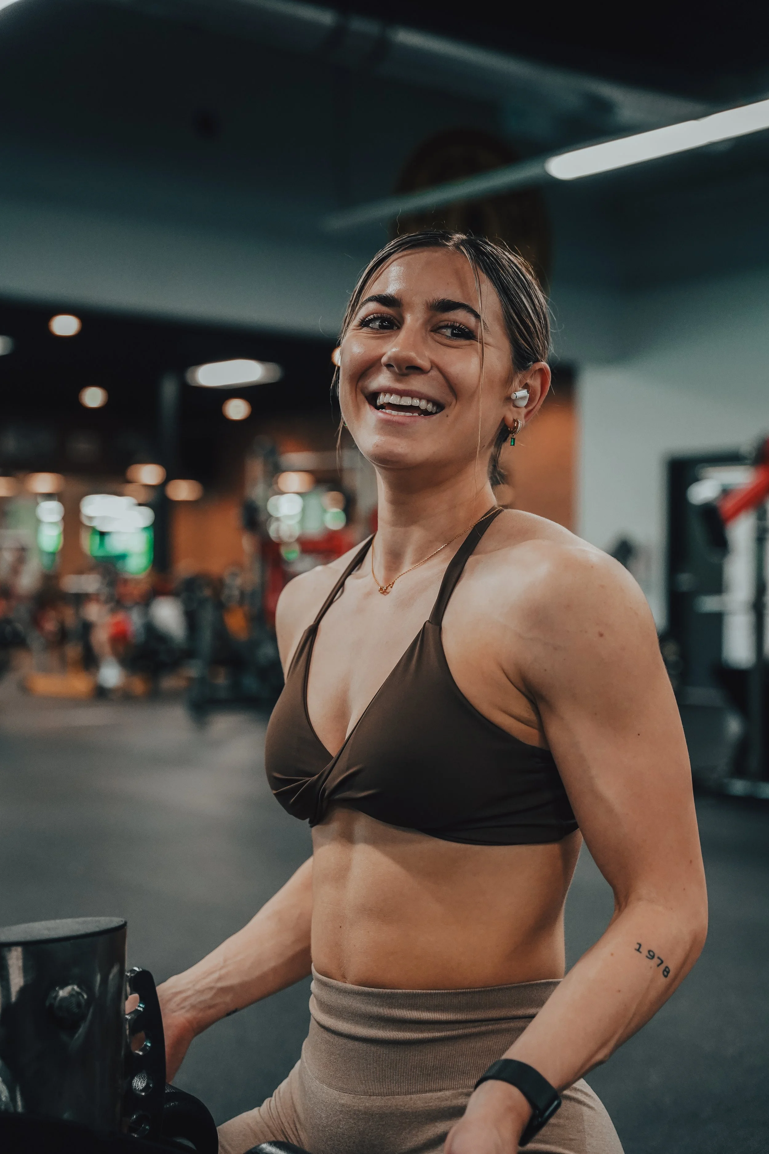 A woman with a tan sports bra and beige workout pants in a gym, smiling and working out with gym equipment.