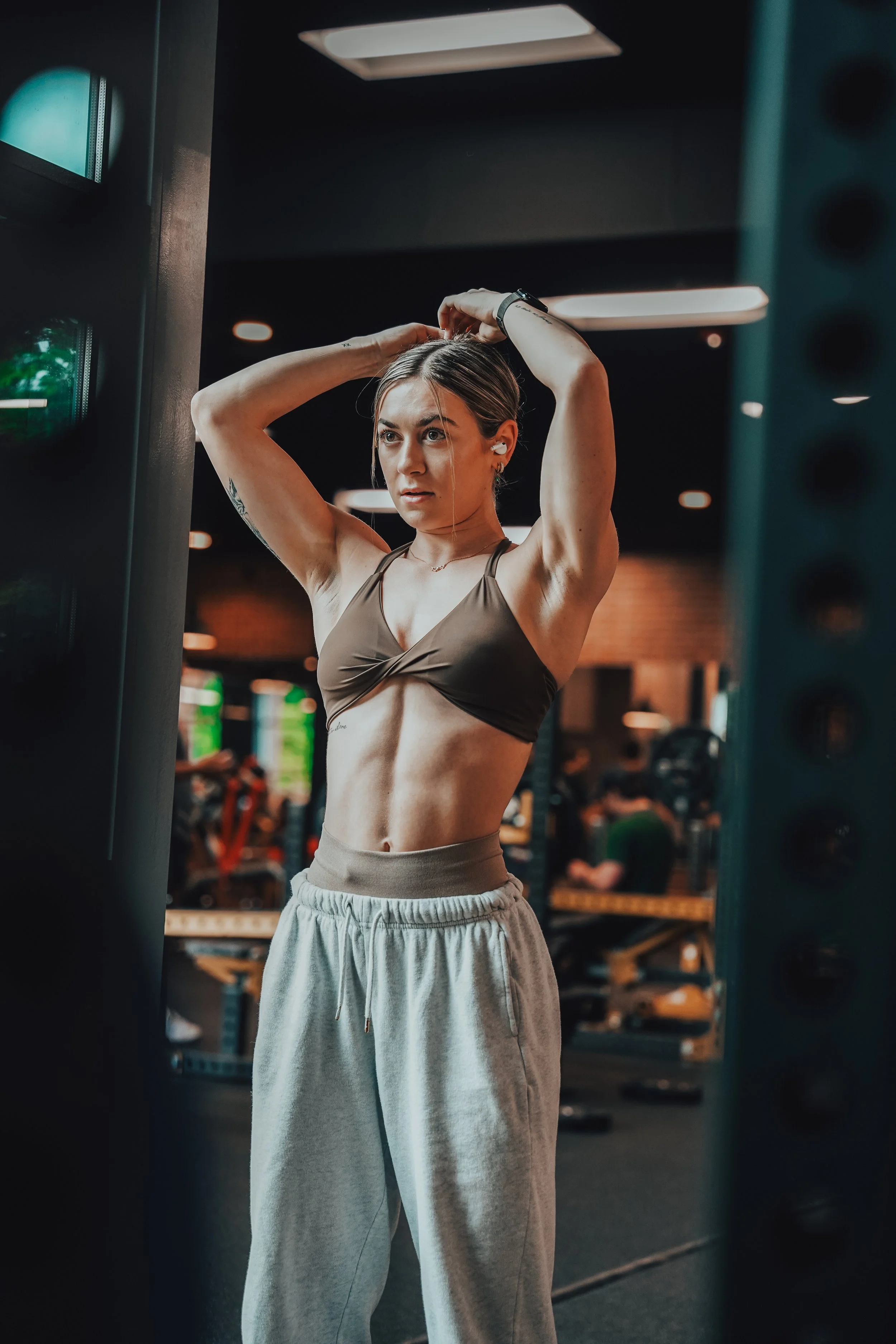 A young woman with tattoos, wearing a brown sports bra and gray sweatpants, is in a gym, adjusting her hair while looking at herself in the mirror.