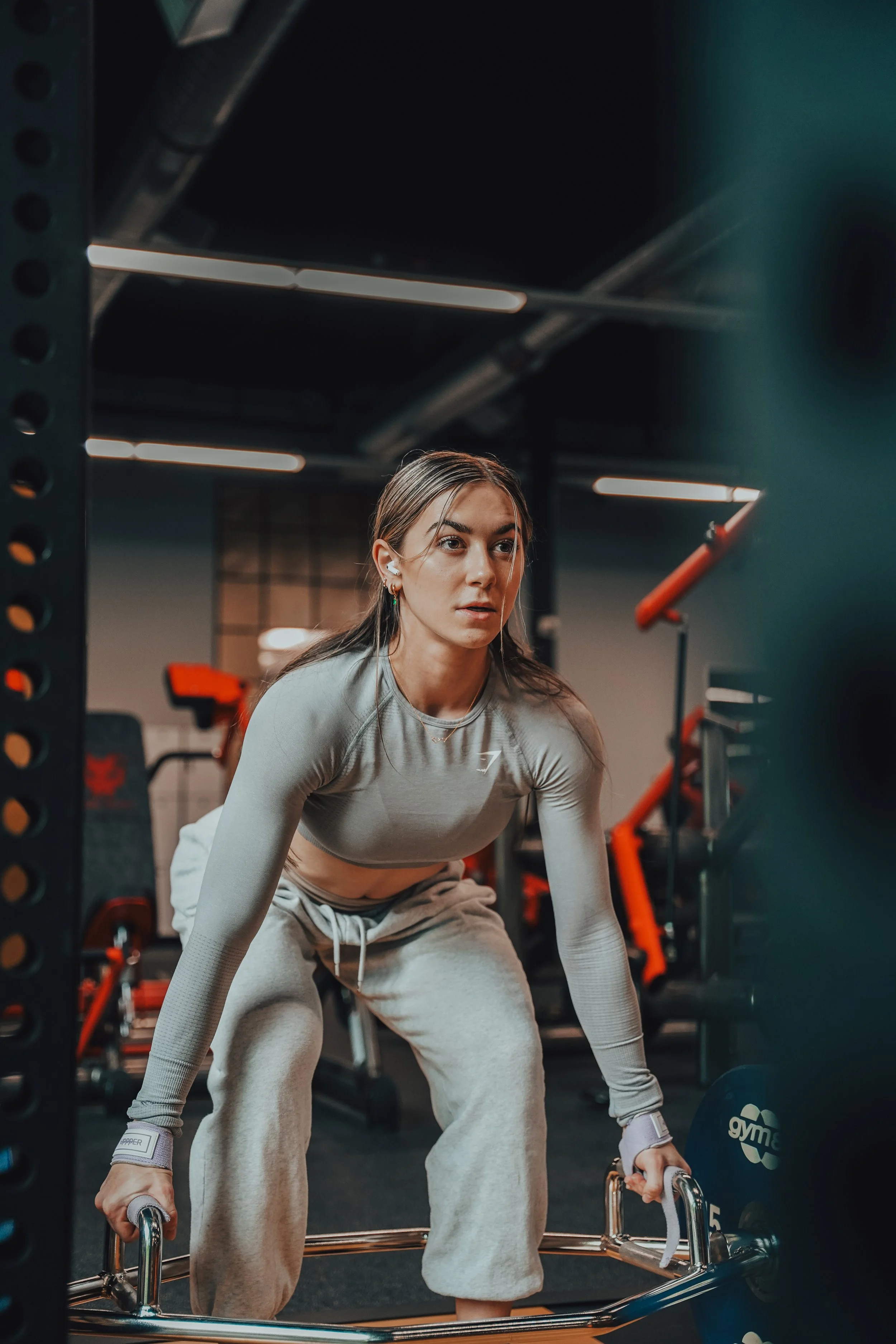 A woman in workout clothing, with wired earbuds, is lifting a barbell in a gym.
