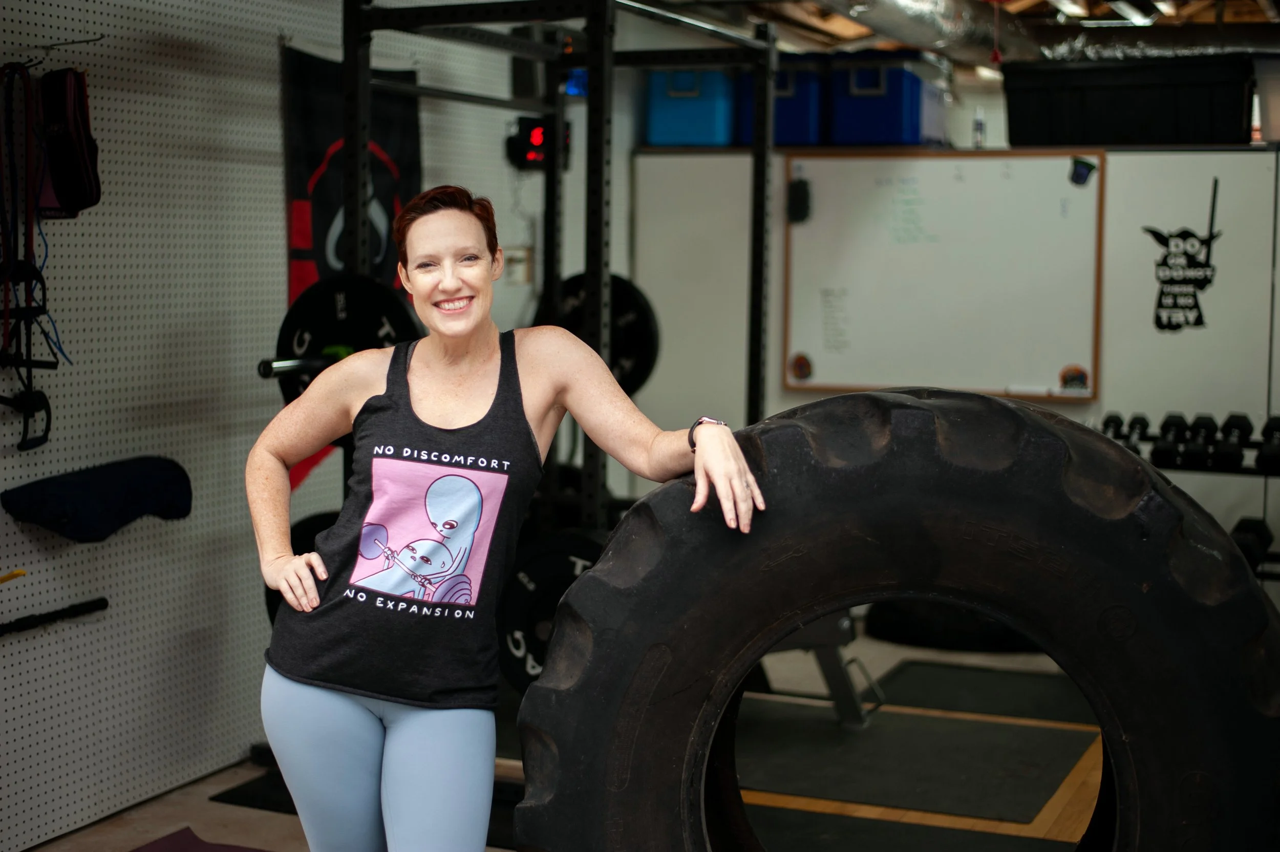 Kristyn McGeehan leaning on a large tire in her home gym wearing a No Discomfort No Expansion shirt