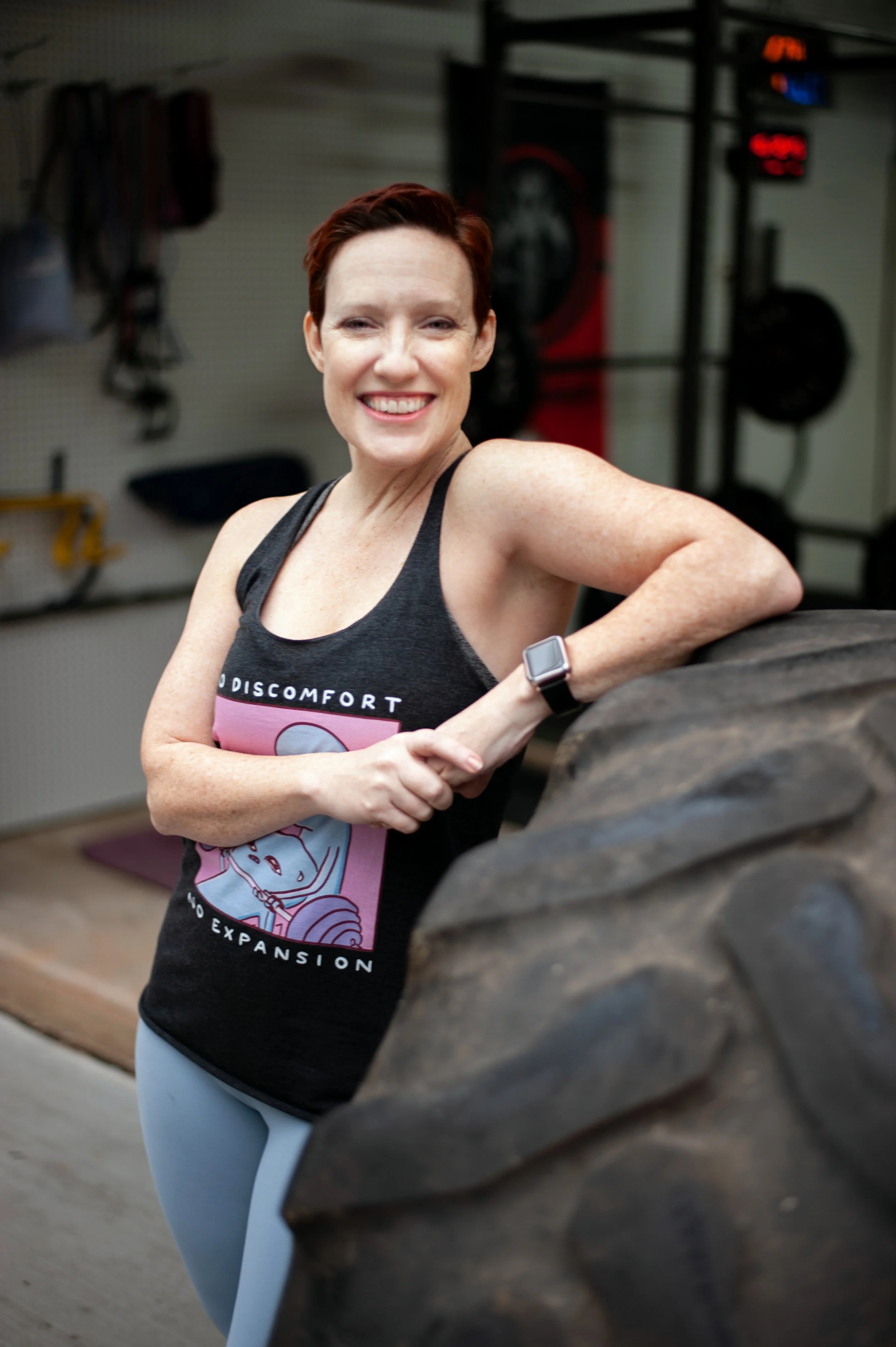 Kristyn McGeehan leaning on a large tire in her home gym wearing a No Discomfort No Expansion shirt