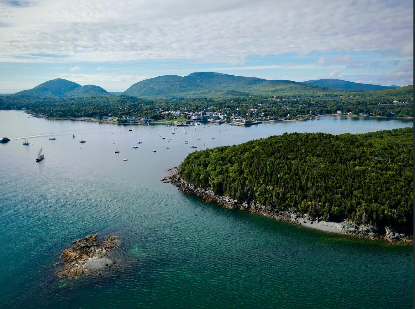 Aerial view of a coastal town with sailboats on the water, green forested islands, and mountains in the background under a partly cloudy sky.
