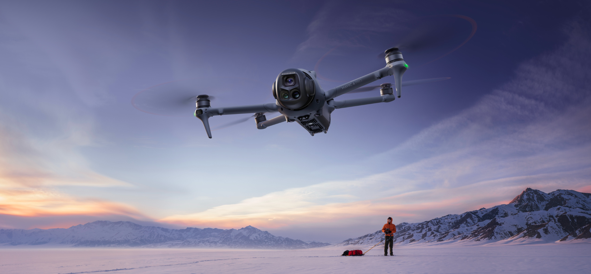 A drone flying over a snowy landscape with mountains in the background and a person standing on the snow holding a leash attached to a sled.