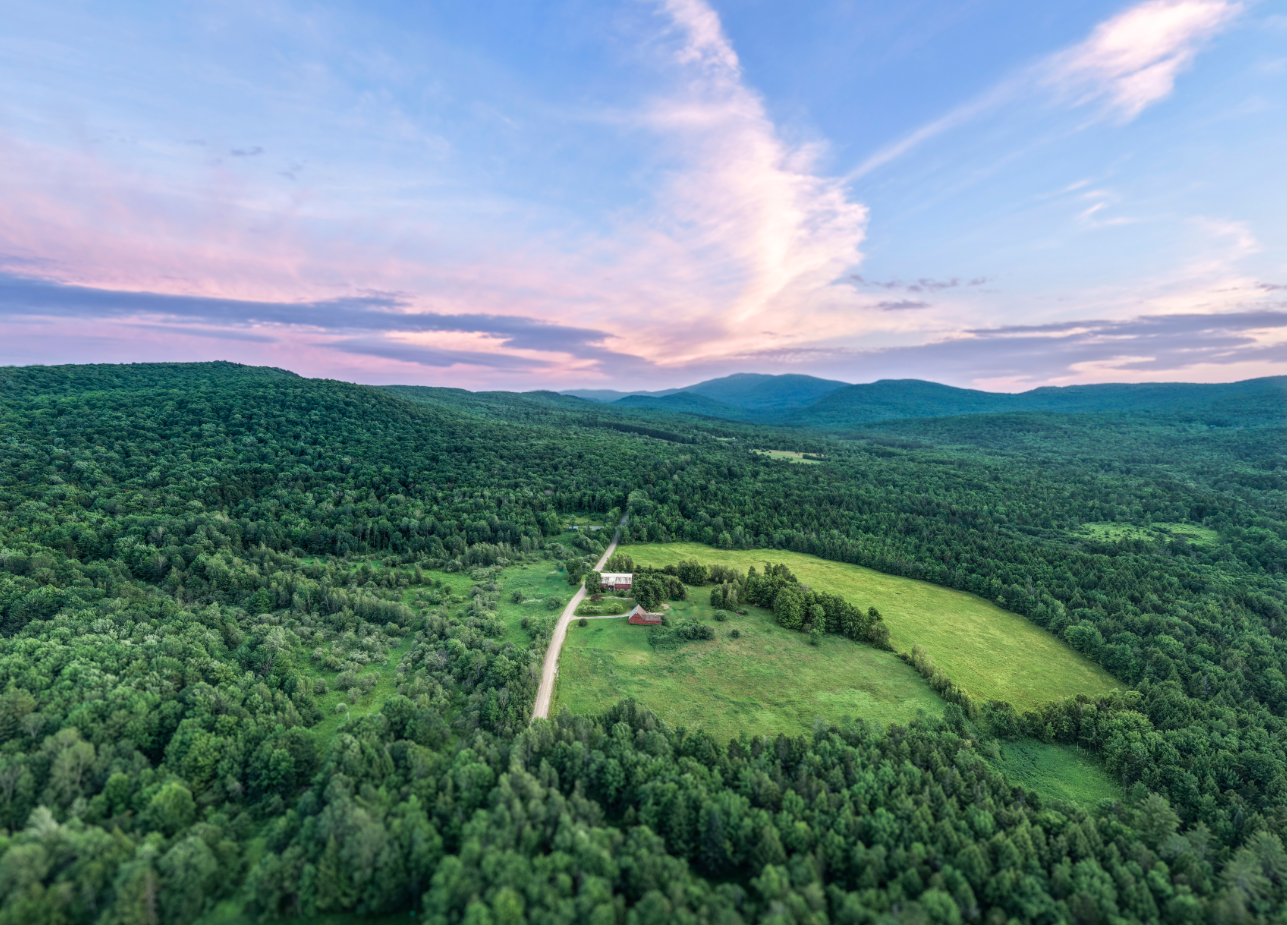 Aerial view of a rural landscape featuring green forests, a small road leading to farmhouses, open fields, and distant mountains under a partly cloudy sky at sunset.