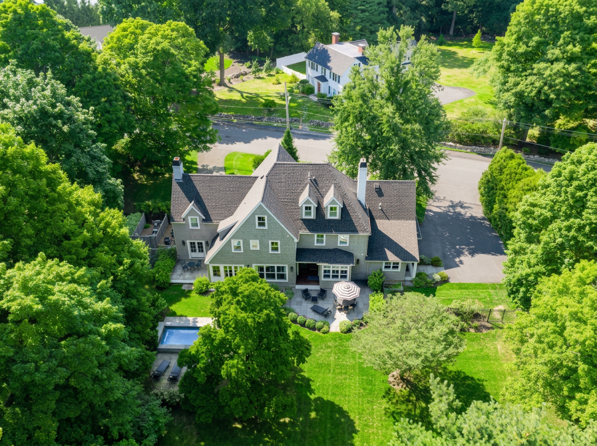 A large, multi-story house with a dark roof, surrounded by lush green trees and a well-maintained lawn. The backyard includes a patio with outdoor furniture, a pool, and a garden.
