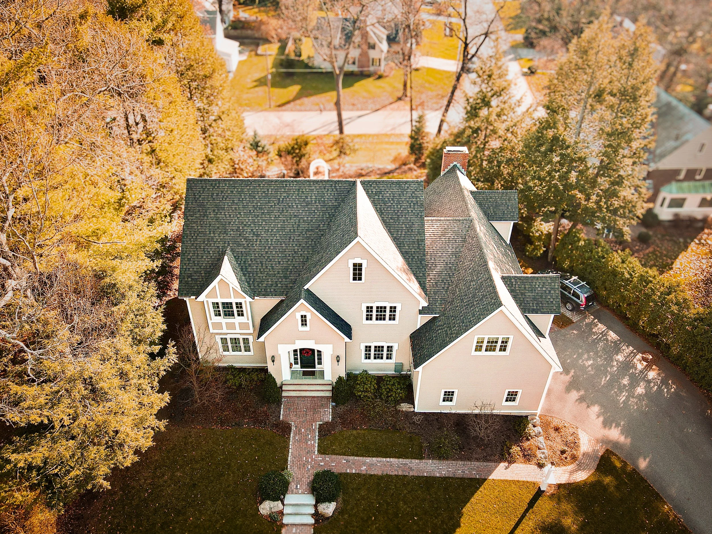 An aerial view of a large house with a gray roof and a brick walkway leading to the front porch, surrounded by fall foliage with trees in shades of yellow and orange.
