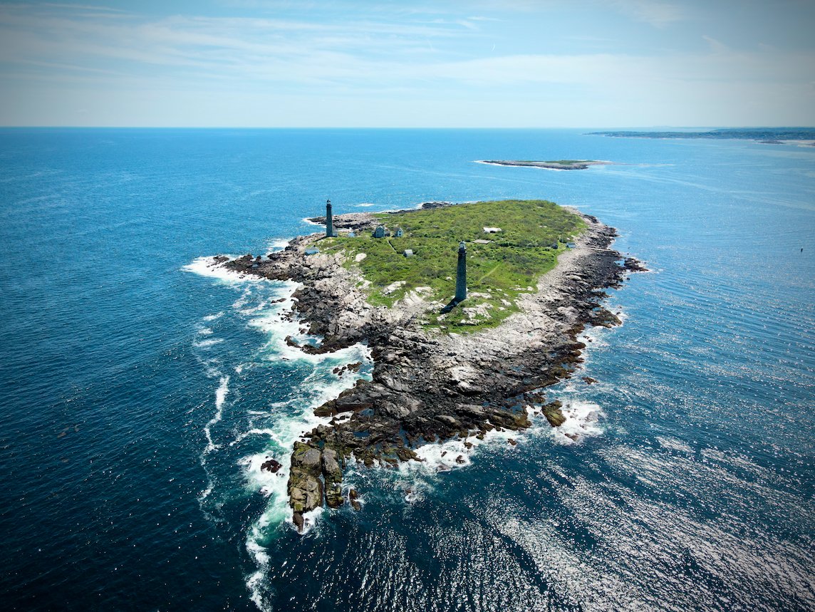 An aerial view of a rocky island with two lighthouses, surrounded by the ocean. The island has green vegetation and a few small buildings, with water crashing against the rocks along the shore.