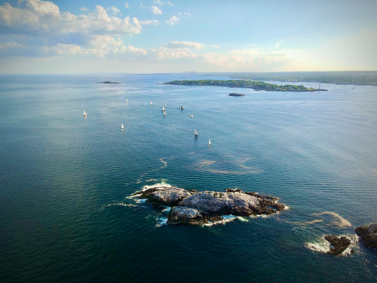 Aerial view of a large body of water with sailboats and small islands, with land and trees in the distance under a partly cloudy sky.