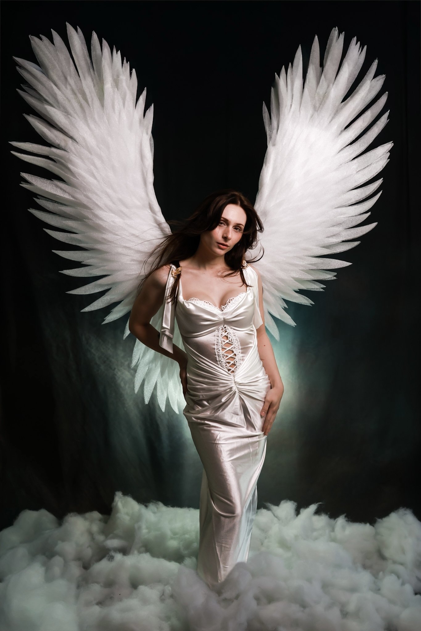 A woman dressed in a silky, white gown with lace-up detail in the front is standing on a fluffy white surface that resembles clouds. She has large, white angel wings behind her and is posing with one hand on her hip, looking at the camera with a conf