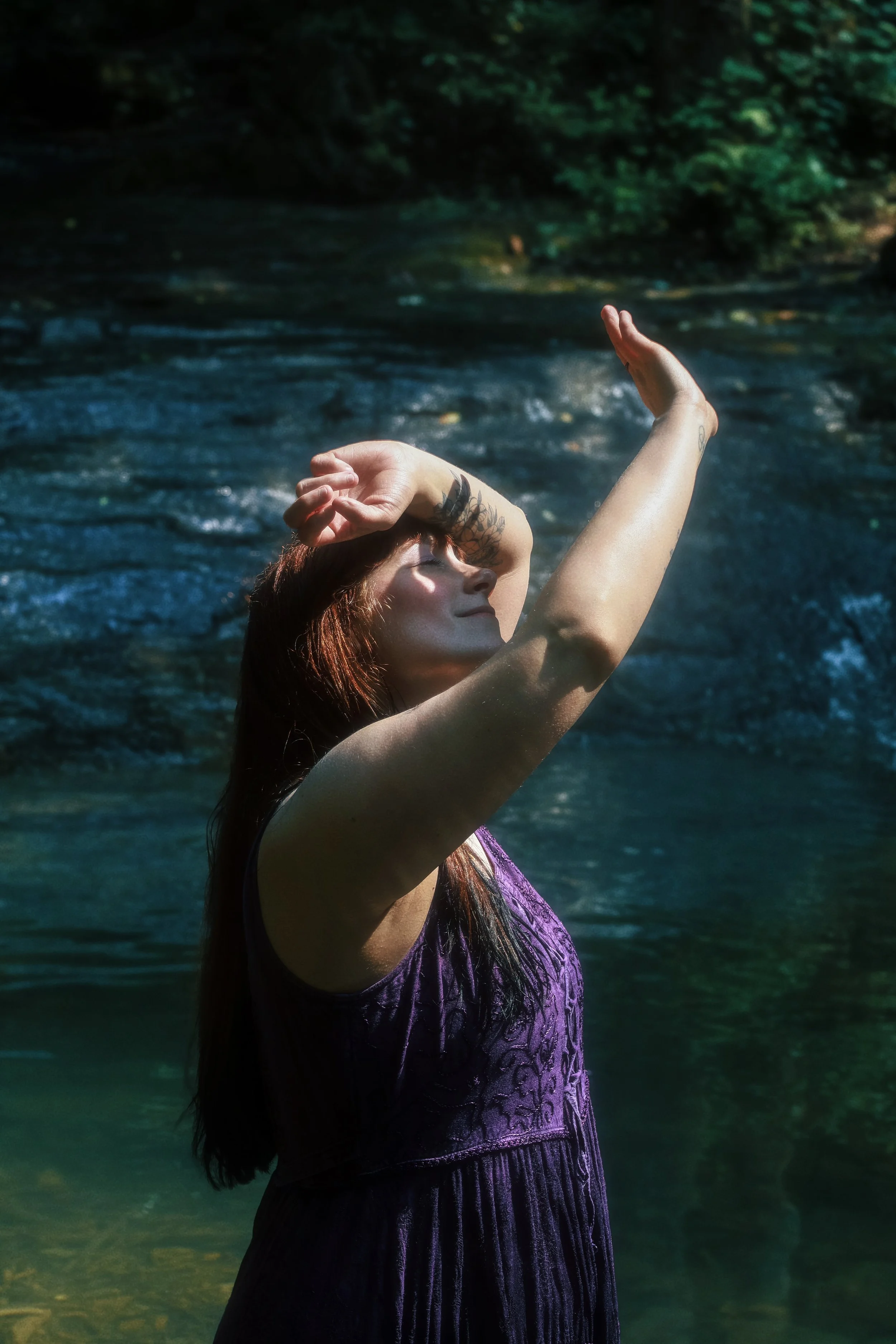 A woman with long hair and tattoos on her arm, standing in a river with her arms raised and eyes closed, surrounded by dense green trees.