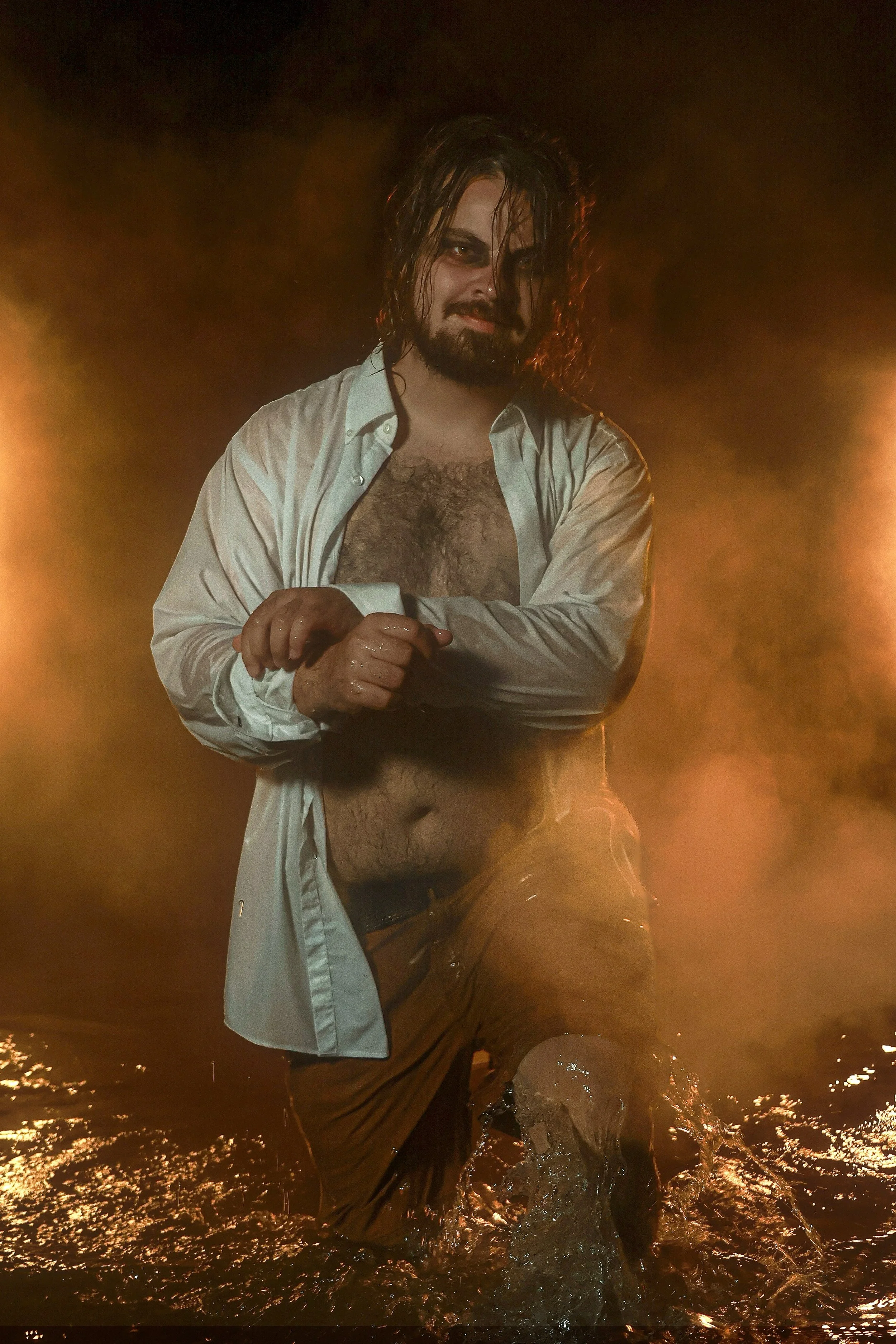 A man with long, wet hair and a beard stands in knee-deep water, wearing an open white shirt and yellow pants, smiling at the camera. The background is dark and illuminated by warm, orange lighting.