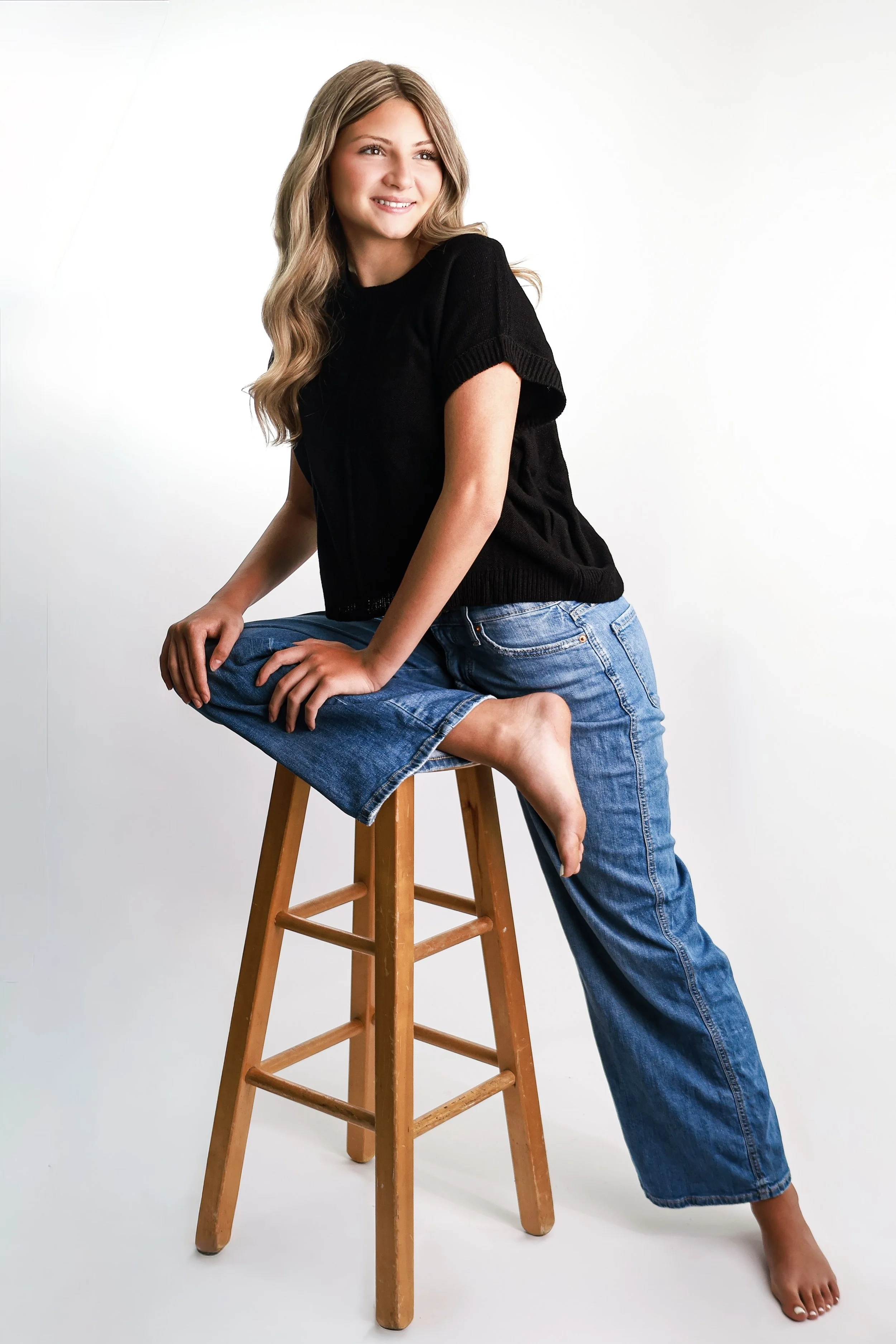A young woman with long wavy blonde hair wearing a black t-shirt and loose blue jeans, sitting on a wooden stool with one leg crossed over the other, smiling and looking slightly to the right against a plain white background.