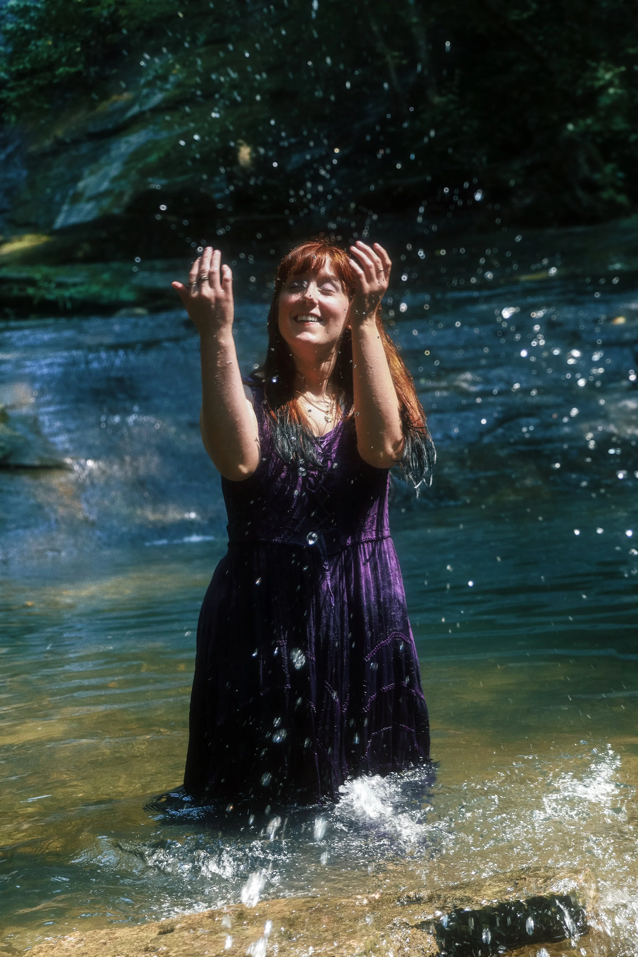 A woman in a purple dress is standing in a river under a rocky overhang, smiling and with her hands raised as water splashes around her.