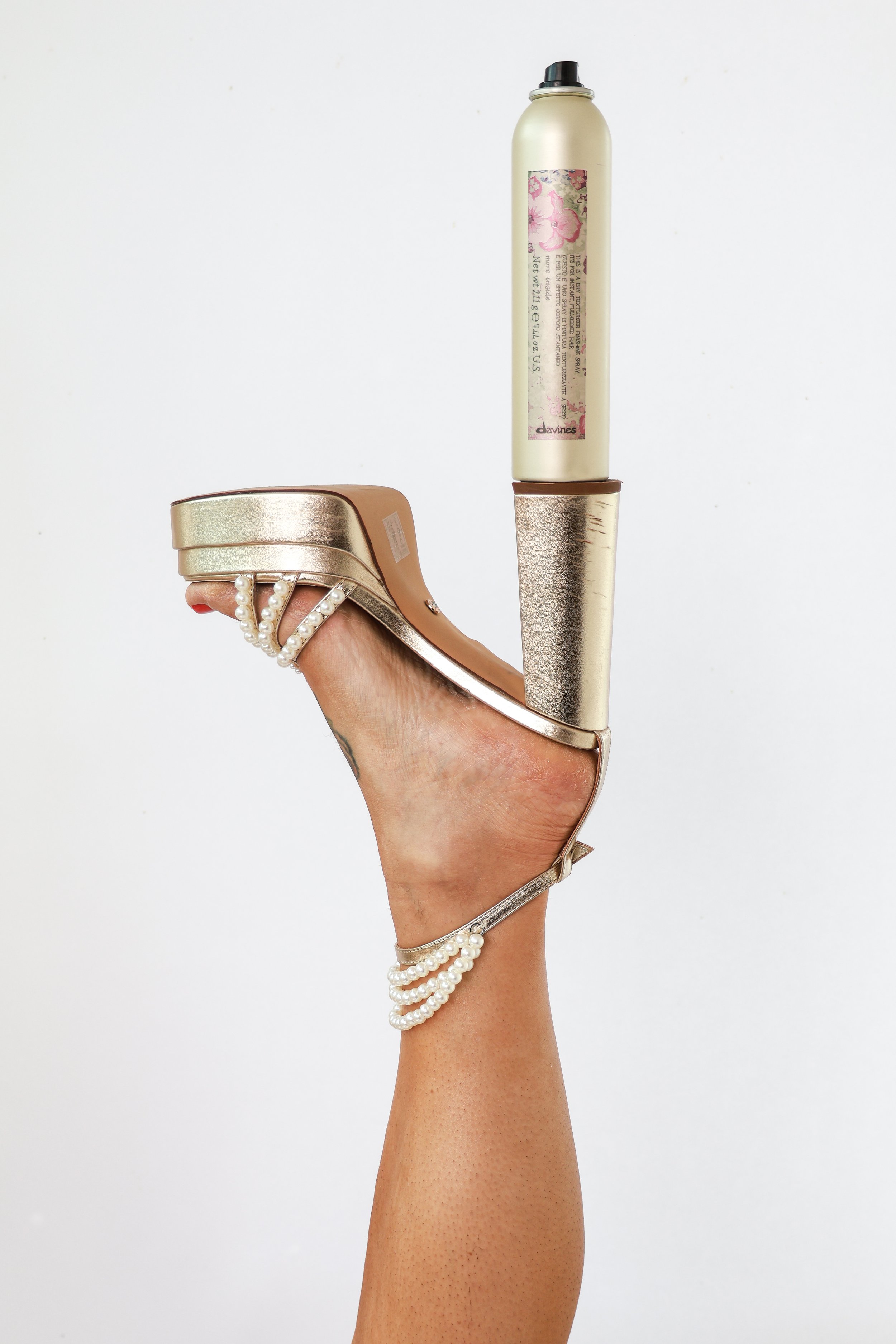A hand with pearl bracelets is holding a high-heel shoe with a gold finish. The shoe has a thick heel and is worn with a gold metallic sock or heel cover. The background is plain white.