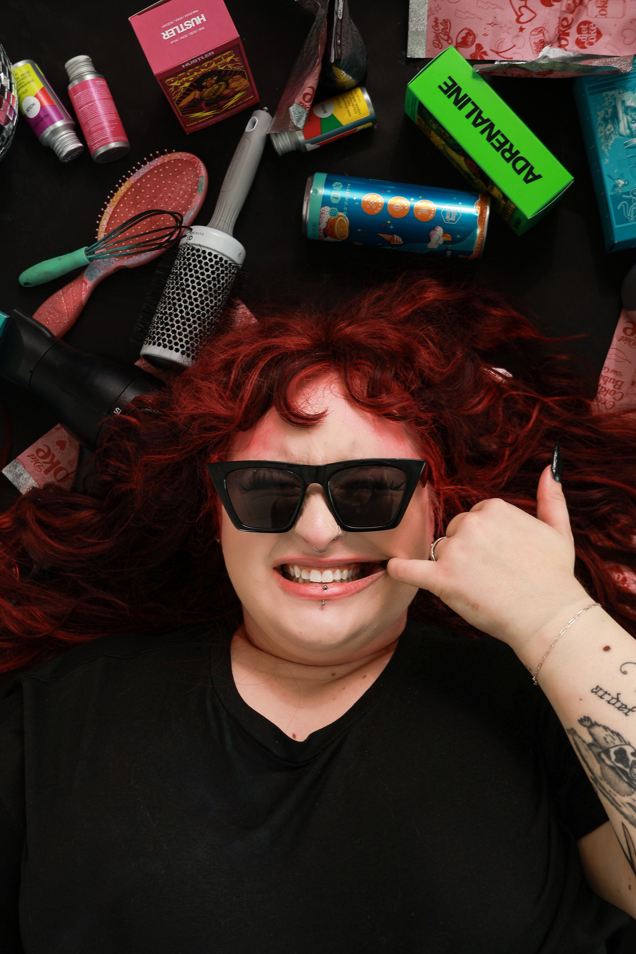 A woman with red curly hair, wearing black sunglasses and a black shirt, lying on her back on a dark surface. She is smiling, showing her teeth, and has a piercing on her lip. She is making a hand gesture near her face with her thumb and pinky extend