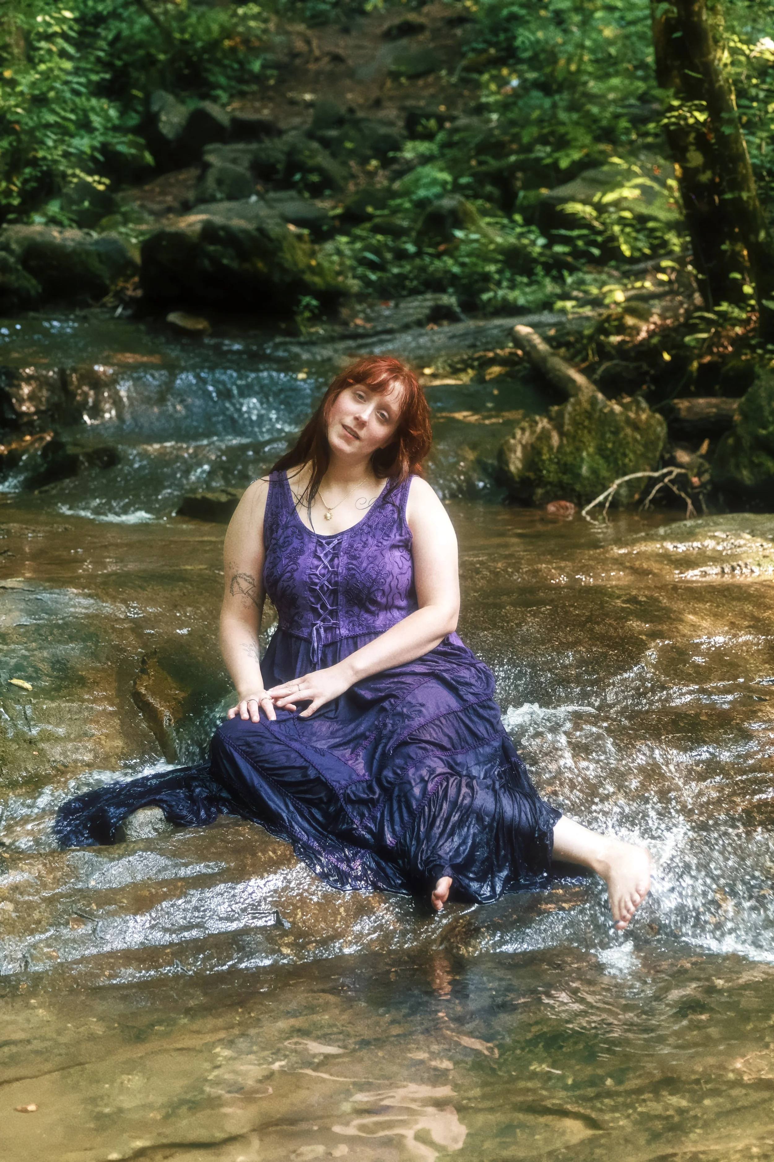 A woman with red hair wearing a purple dress sitting in a shallow stream in a lush green forest.