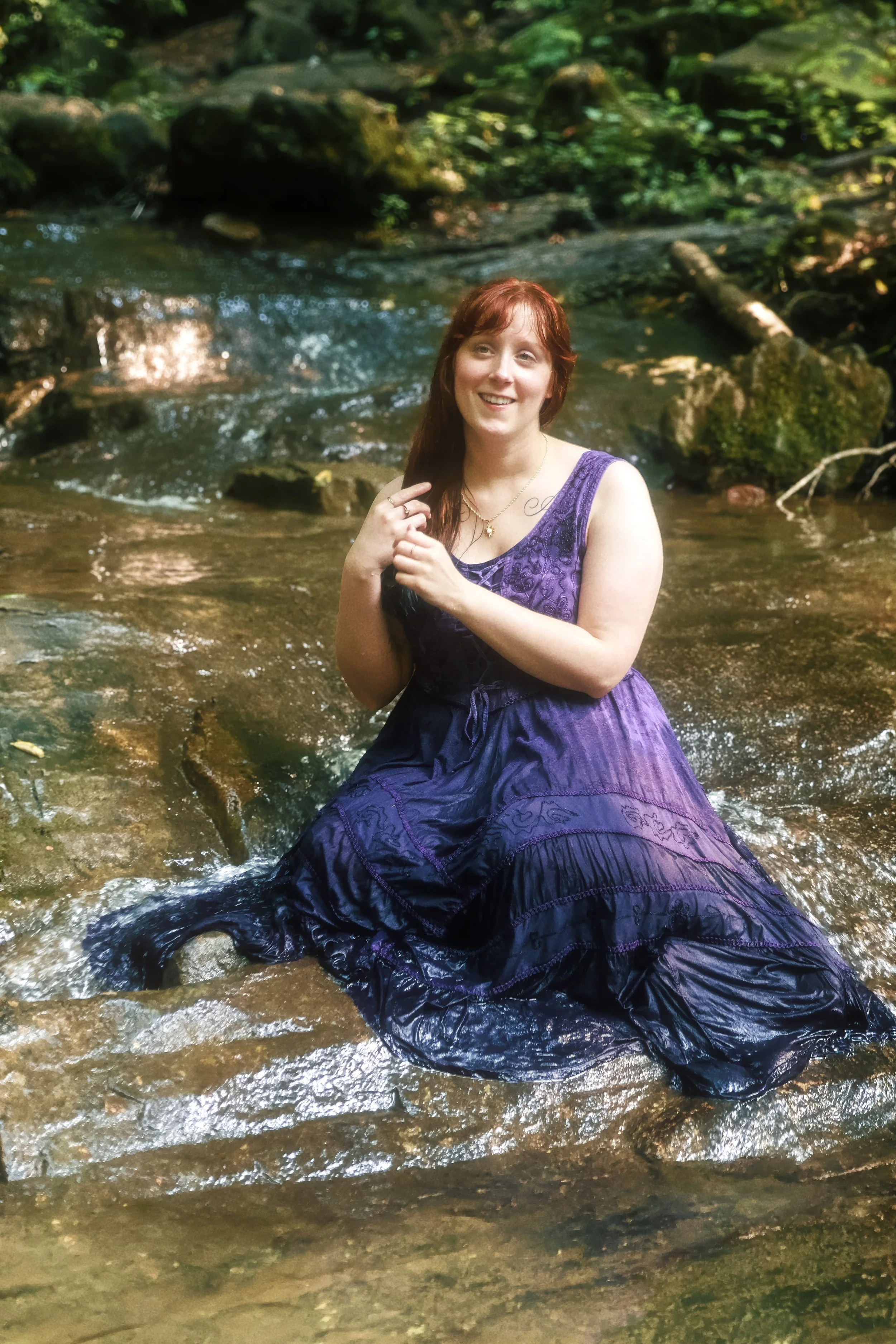 A woman with red hair wearing a purple dress sitting in a shallow creek with green foliage and rocks in the background.
