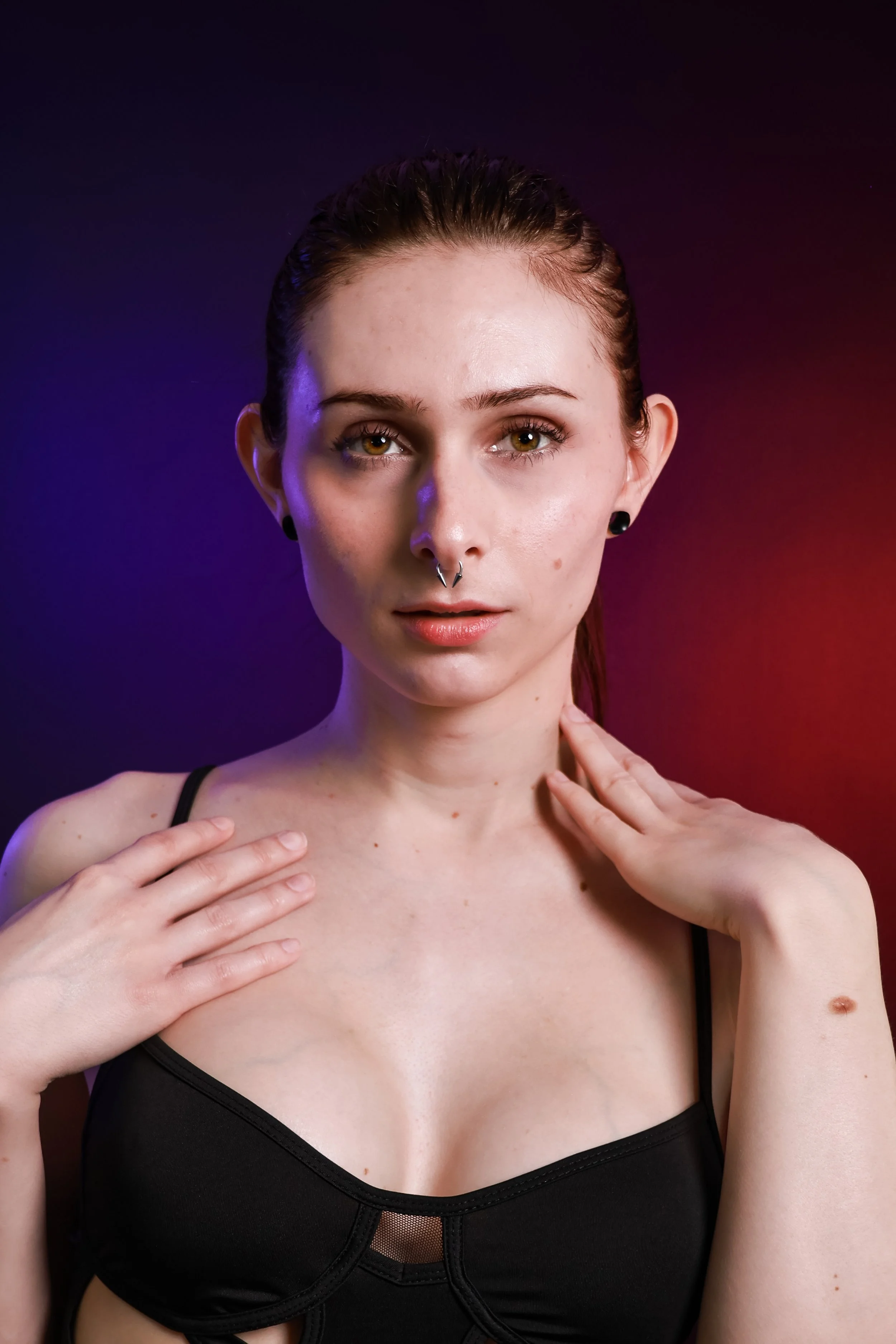 A young woman with brown hair pulled back, wearing a black bra, holds her shoulders with her hands, looking directly at the camera against a dark background illuminated with red and purple lighting.